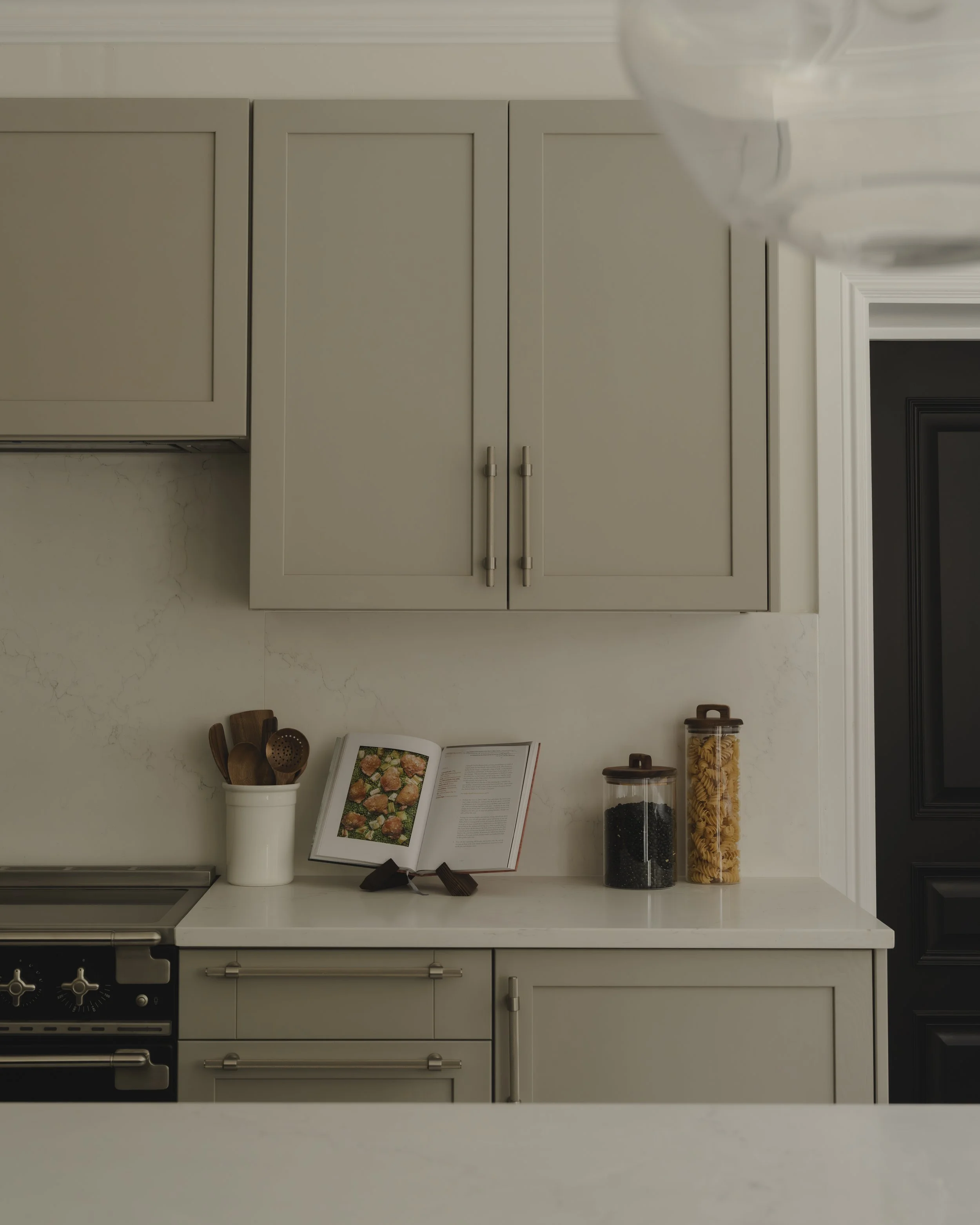 Kitchen countertop with an open cookbook, jars of pasta and spices, utensil holder, and a stovetop below grey cabinets.