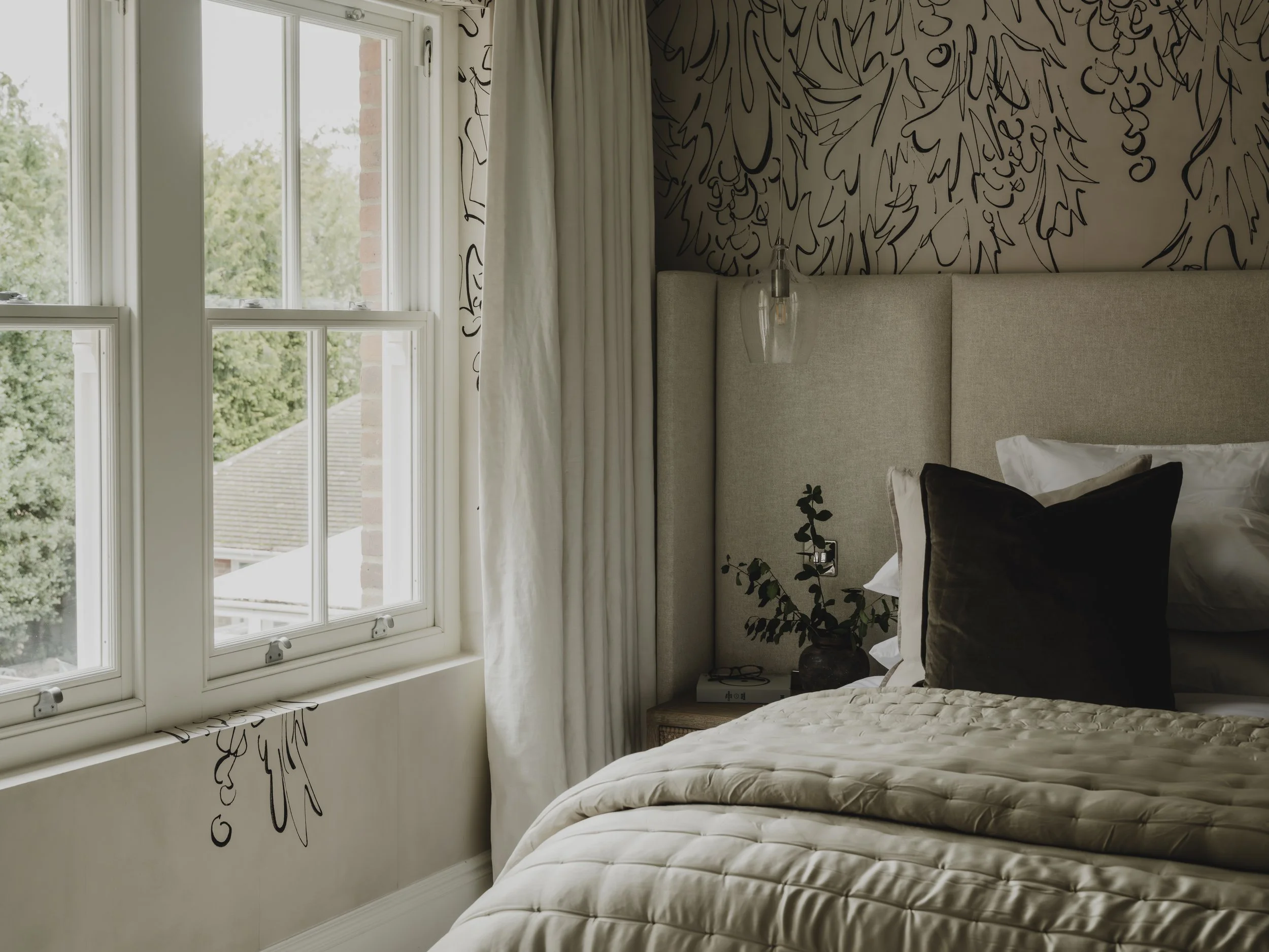A cozy bedroom with a large window, a upholstered bed with white and dark pillows, a bedside table with a plant, and decorative black and white wall art.