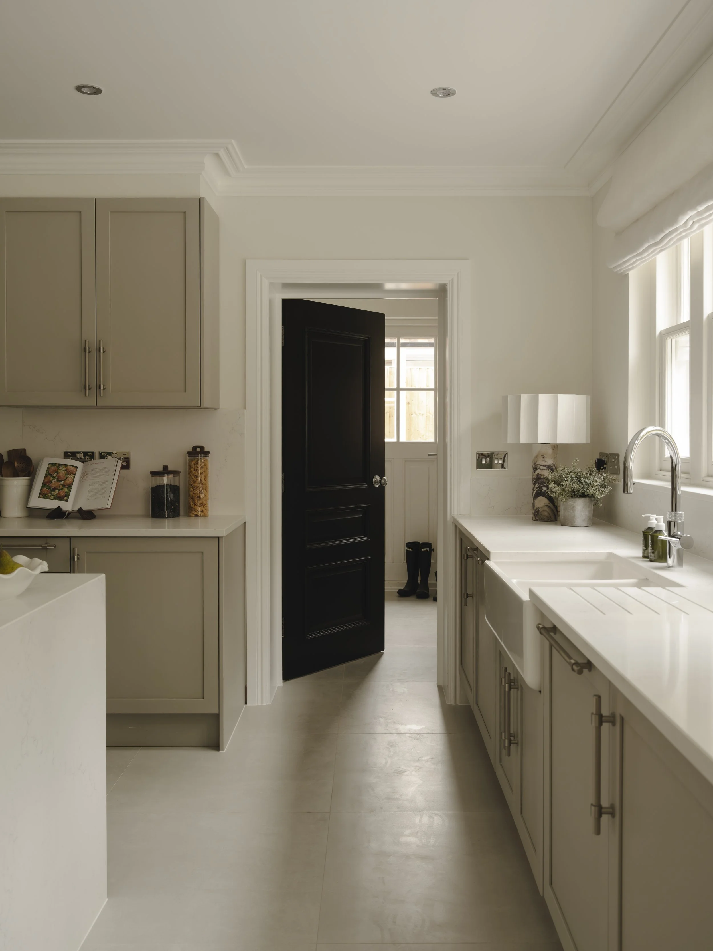 A kitchen with beige cabinets, a white countertop, and a black partially open door leading to a mudroom or back entrance with a window. There are two small jars of pasta and spices on the counter, a book open on a stand, and a lamp with a vase of flo