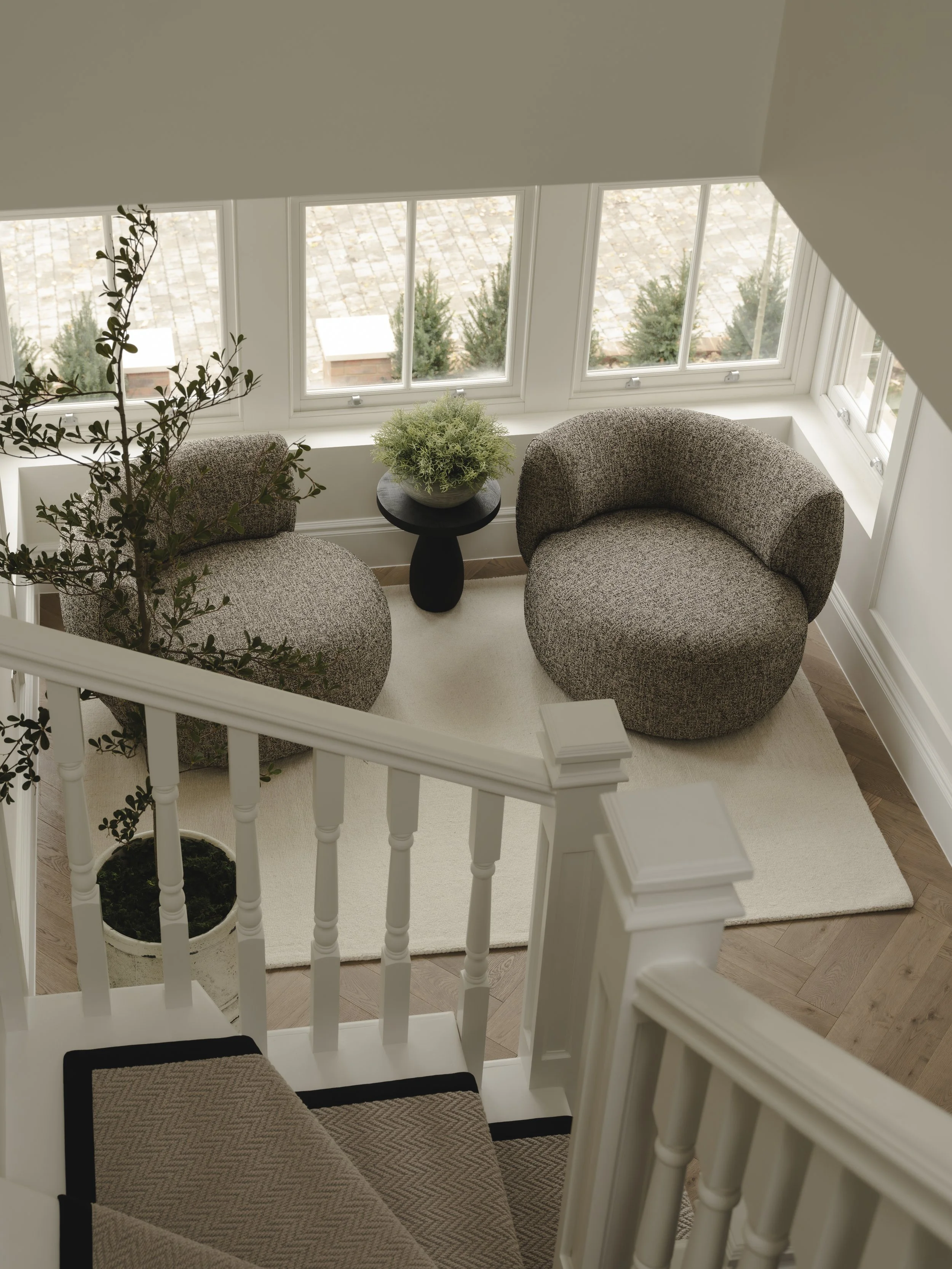 A cozy sitting area with two rounded, upholstered armchairs in a light brown and gray fabric, a small black side table with a green plant, a white rug, and a window seat at the top of a staircase with a beige and black patterned cushion.