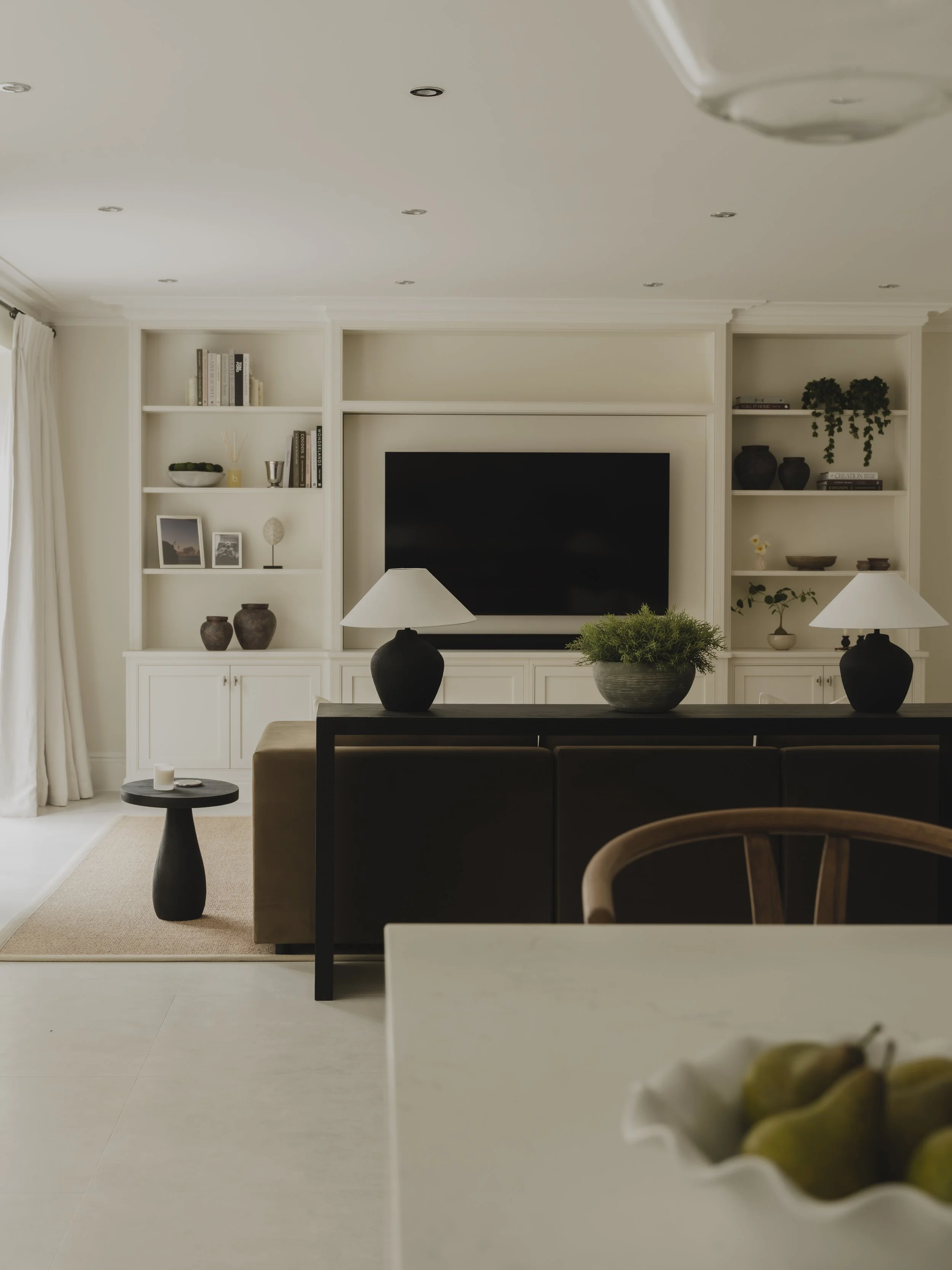 Living room with built-in white shelves, a large TV, beige sofa, black side tables, lamps, and decorative items including plants and vases.