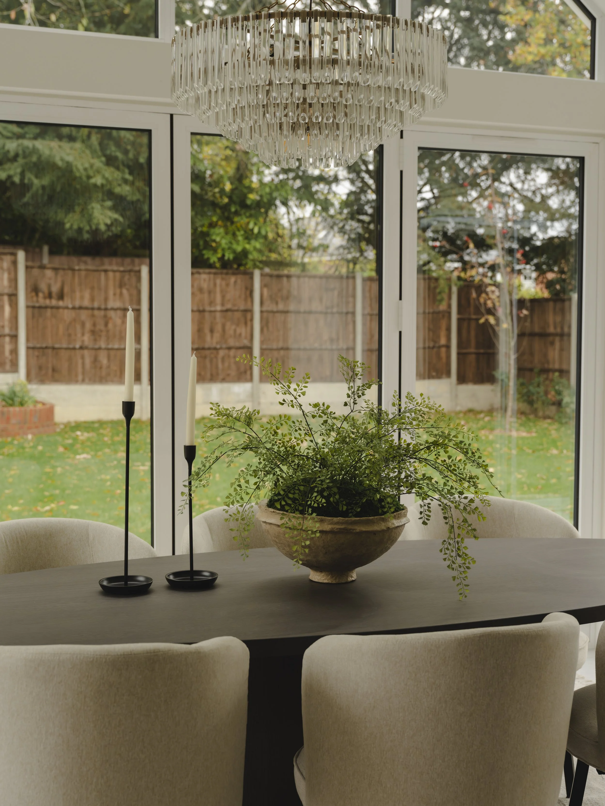 A dining room table with a potted green plant and two black candle holders with white candles, situated in front of large glass windows with a view of a fenced backyard with trees and green grass, illuminated by a chandelier hanging from the ceiling.