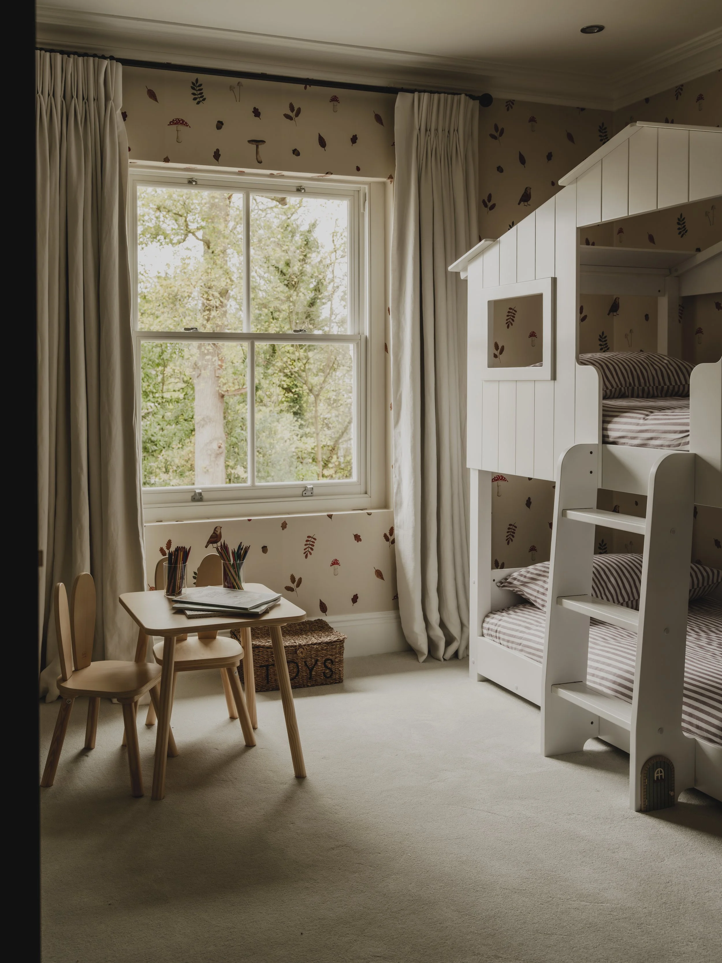 Child's bedroom with a window, white bunk bed, small wooden desk with chairs, and a basket labeled 'TOYS' in a cozy, decorated room.