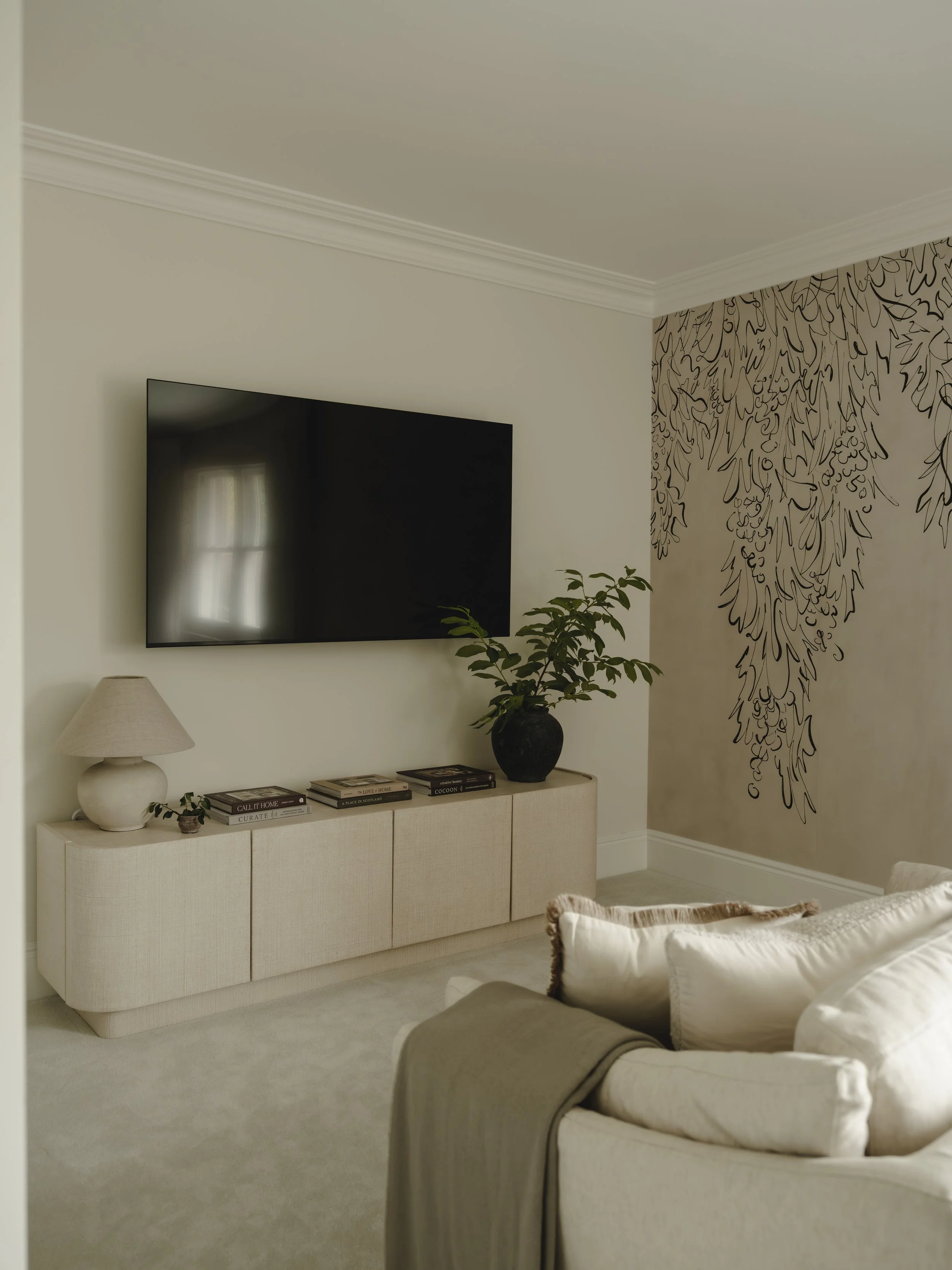 Living room with a wall-mounted flat-screen TV, beige console table with books, a white lamp, and a black vase with green leaves. There is a cream-colored sofa with pillows in the foreground, and a decorative black line drawing on the wall to the rig