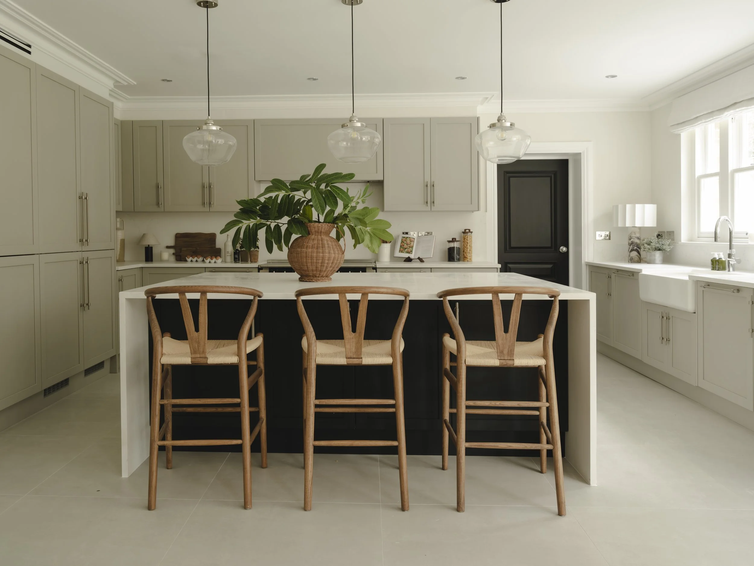 Modern kitchen with light gray cabinets, a white kitchen island with a large potted green plant, and three wooden bar stools. There are pendant lights hanging above the island, a black door, and a window with white trim.