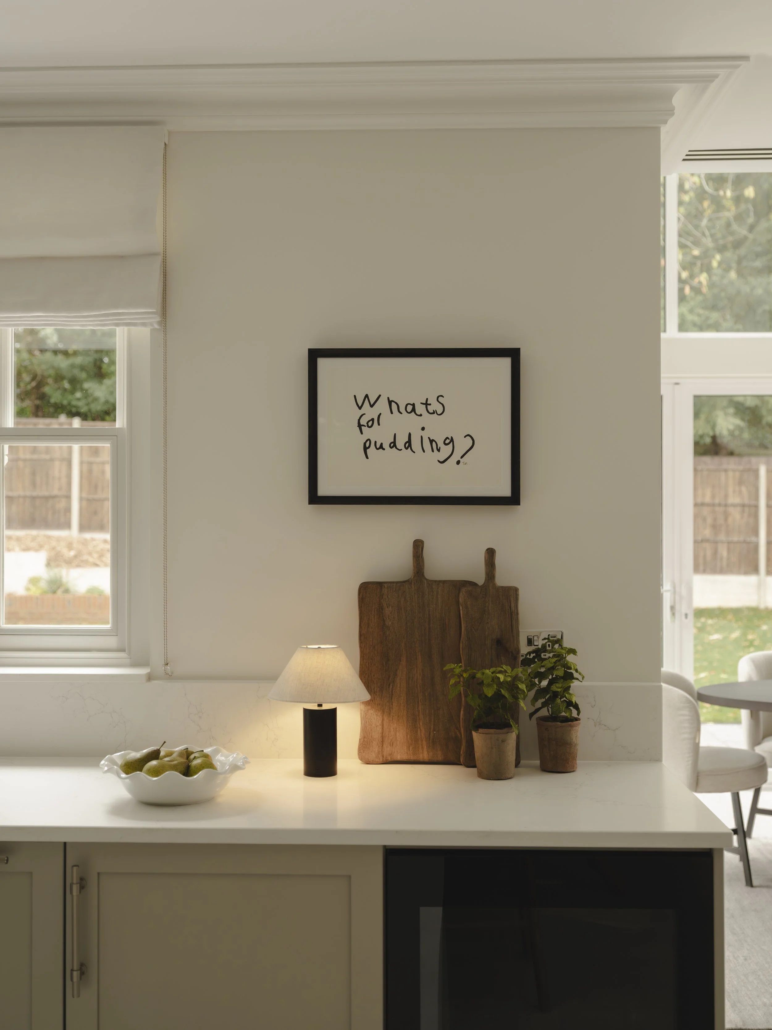 A kitchen counter with a bowl of pears, two wooden cutting boards, a lamp, and potted plants. Framed blackboard on the wall reads 'Whats for pudding?'. Windows in the background reveal a backyard with a wooden fence.