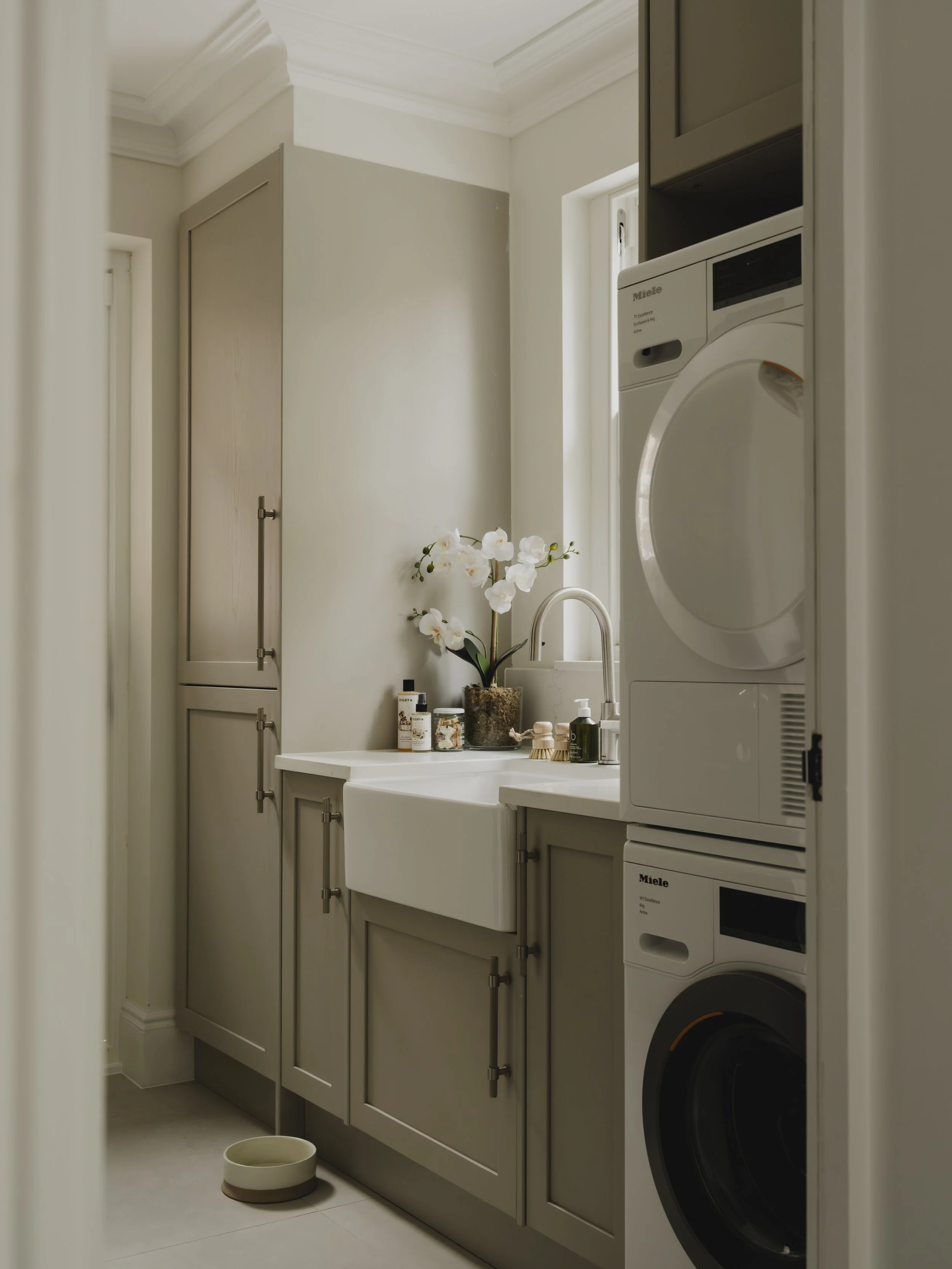 A small laundry room with beige cabinetry, a white farmhouse sink, and a stack of Miele washing machines and dryers. Decor includes a potted orchid and laundry supplies on the counter.