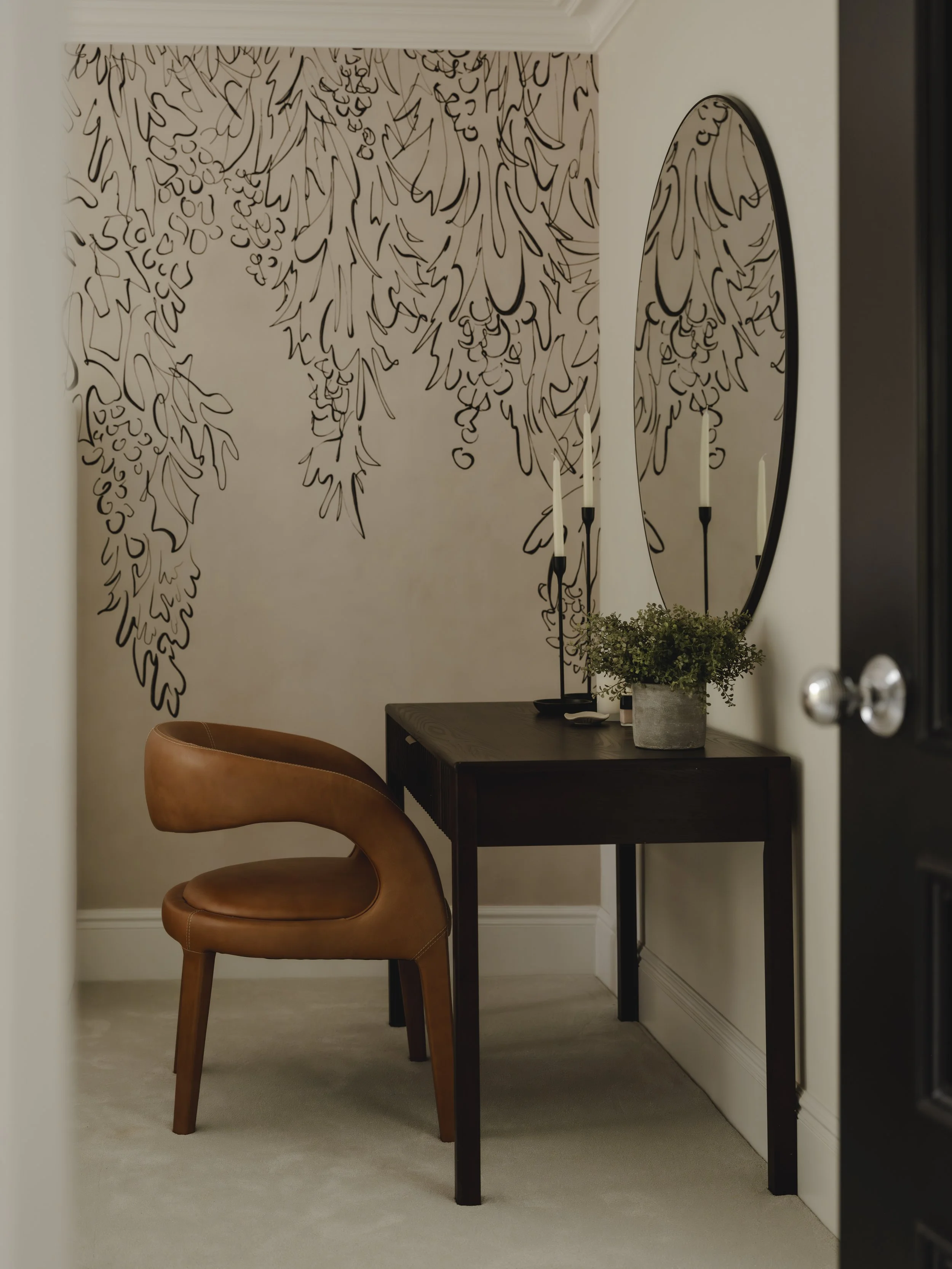 A small interior space with a dark wooden desk, a brown leather chair, and an oval mirror on the wall. Two tall white candles in black candleholders and a potted green plant are on the desk. The wall behind has a black abstract line drawing mural.