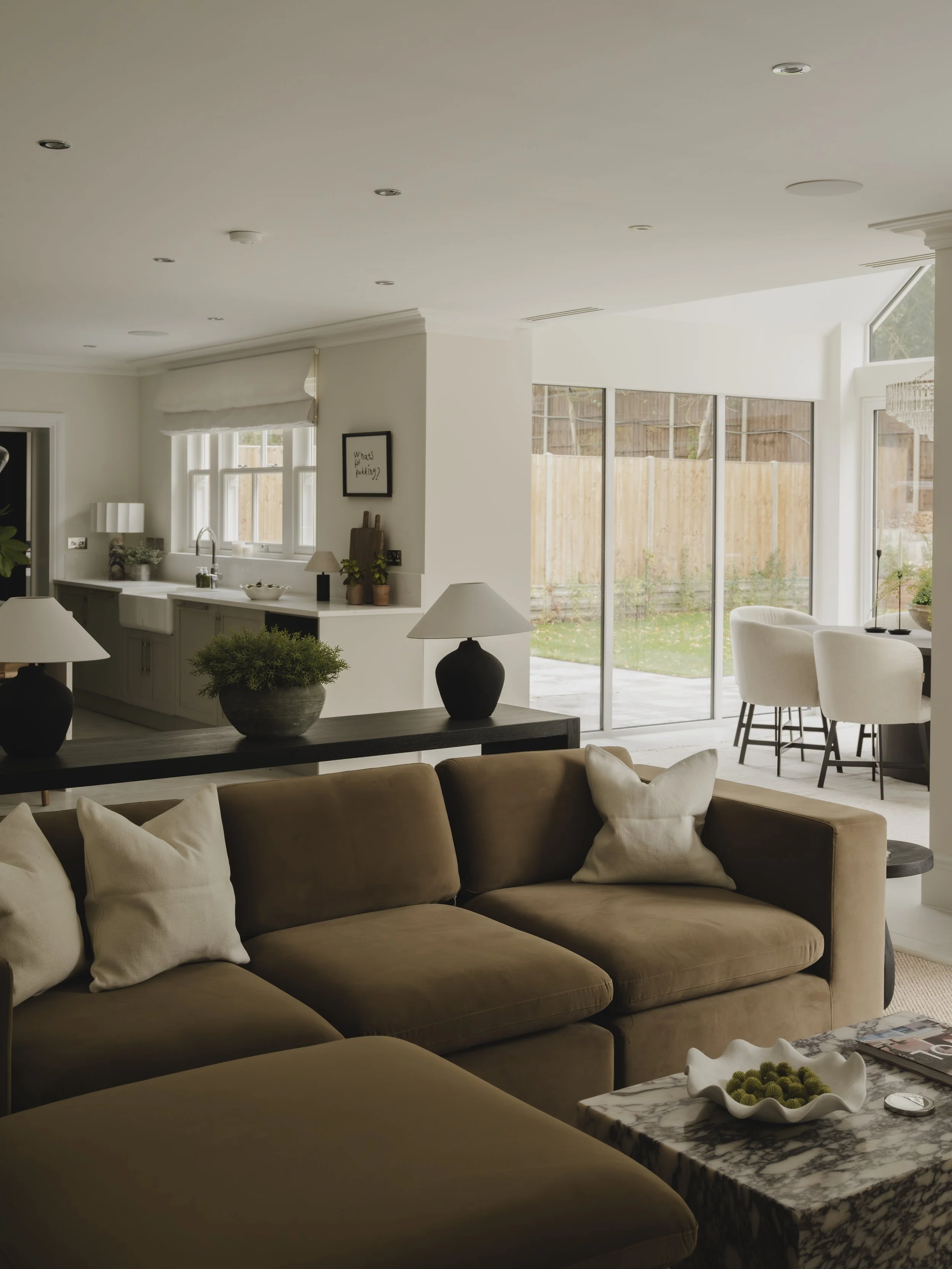 Interior of a living room with a beige sectional sofa, white pillows, dark wood side table with a potted plant, marble coffee table with a decorative bowl, and an open kitchen and dining area with large windows and sliding glass doors in the backgrou