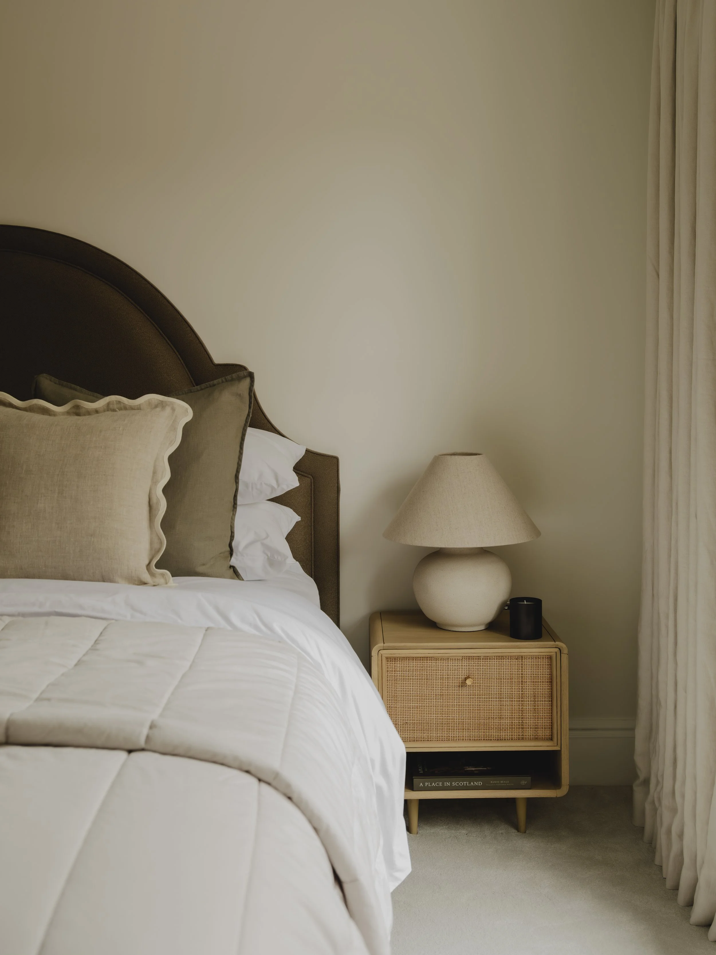 A cozy bedroom corner with a bed topped with beige and white pillows, a wooden nightstand with a ceramic lamp, a black cylindrical object, and a book, next to a window with cream curtains.