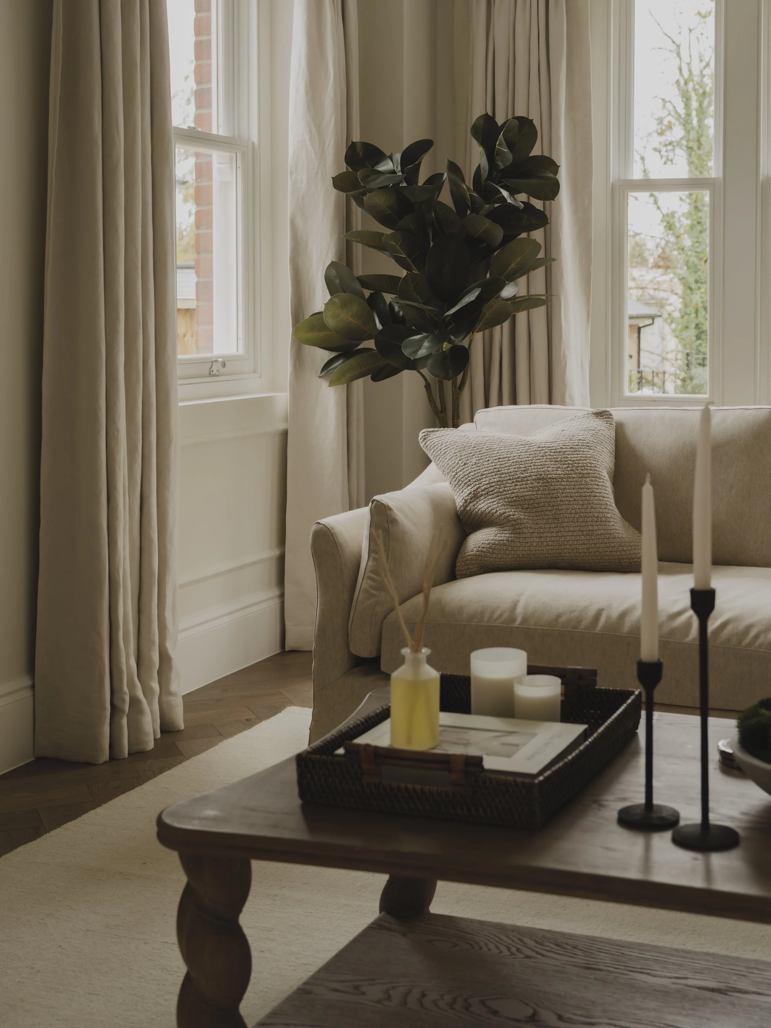 A cozy living room corner with a beige sofa, textured pillows, a large potted plant, and a wooden coffee table with candles and decor, near cream curtains and windows.