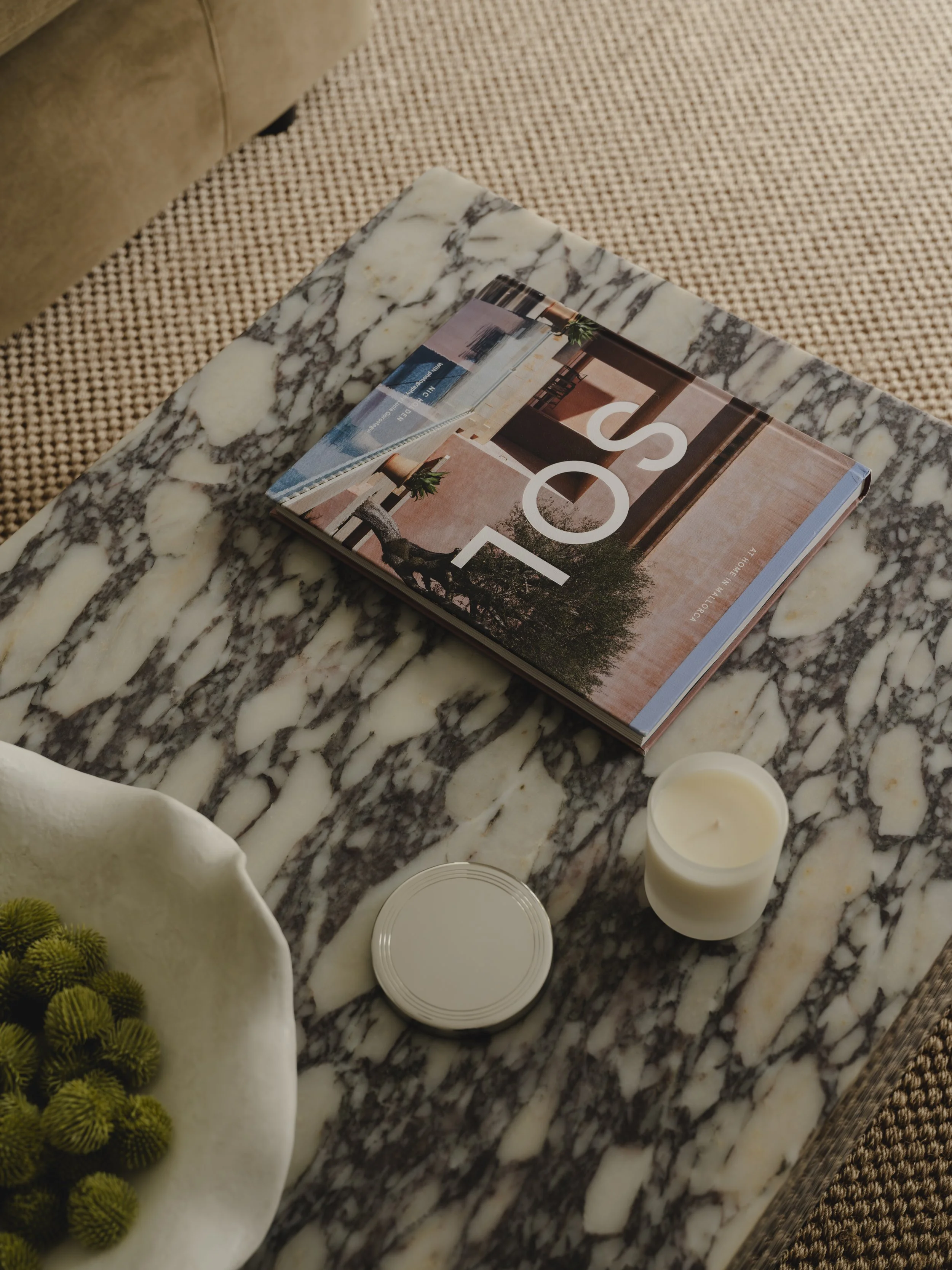 A marble table with a magazine, a small candle in a white jar, a metal tin, and a white bowl with green flowers inside.
