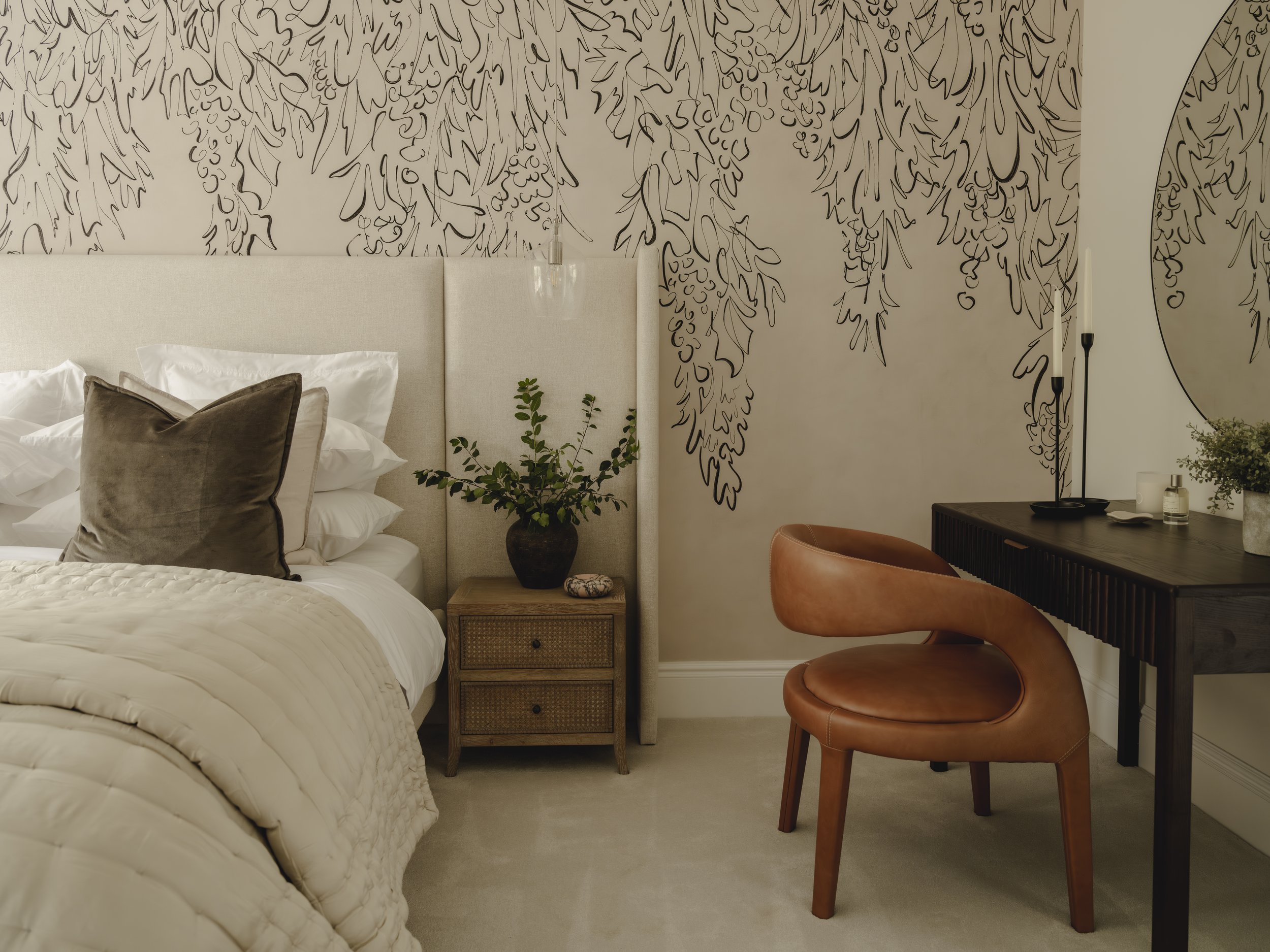 A bedroom with a bed on the left, topped with white and gray pillows, next to a nightstand with a plant. To the right, there's a dark desk with a tan leather chair, a round mirror, and decorative items on the desk. The wall behind features a black li