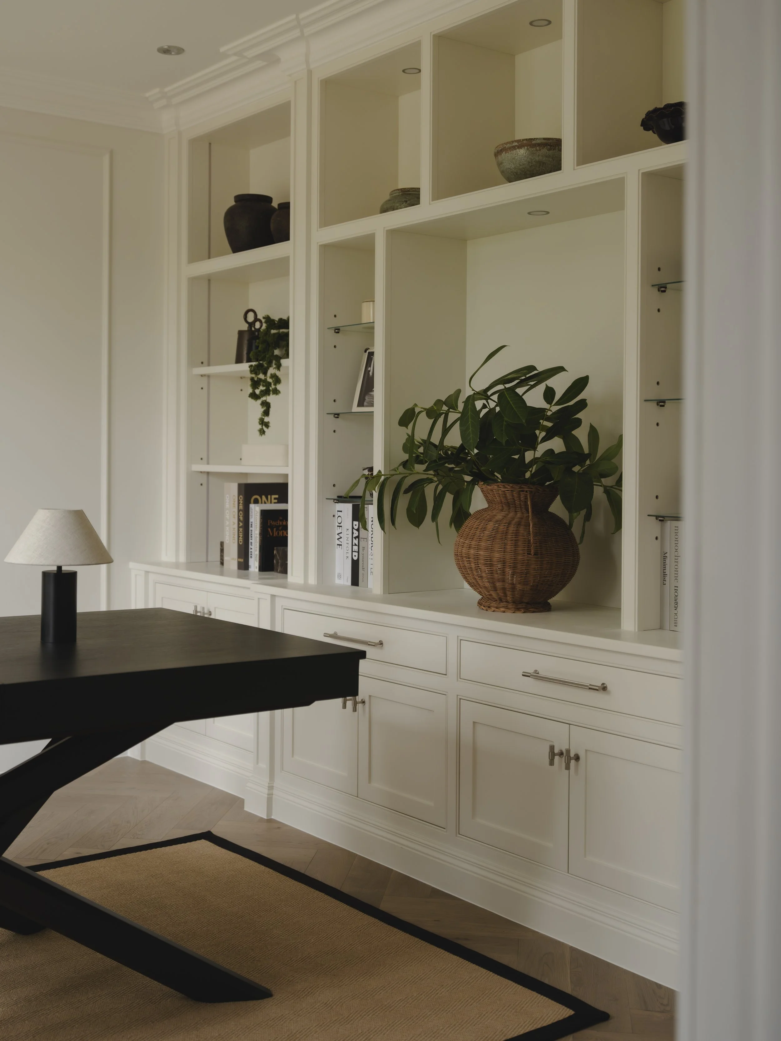 A white built-in bookshelf with plants, books, and decorative items inside a room with a wooden floor and a black table and beige rug.
