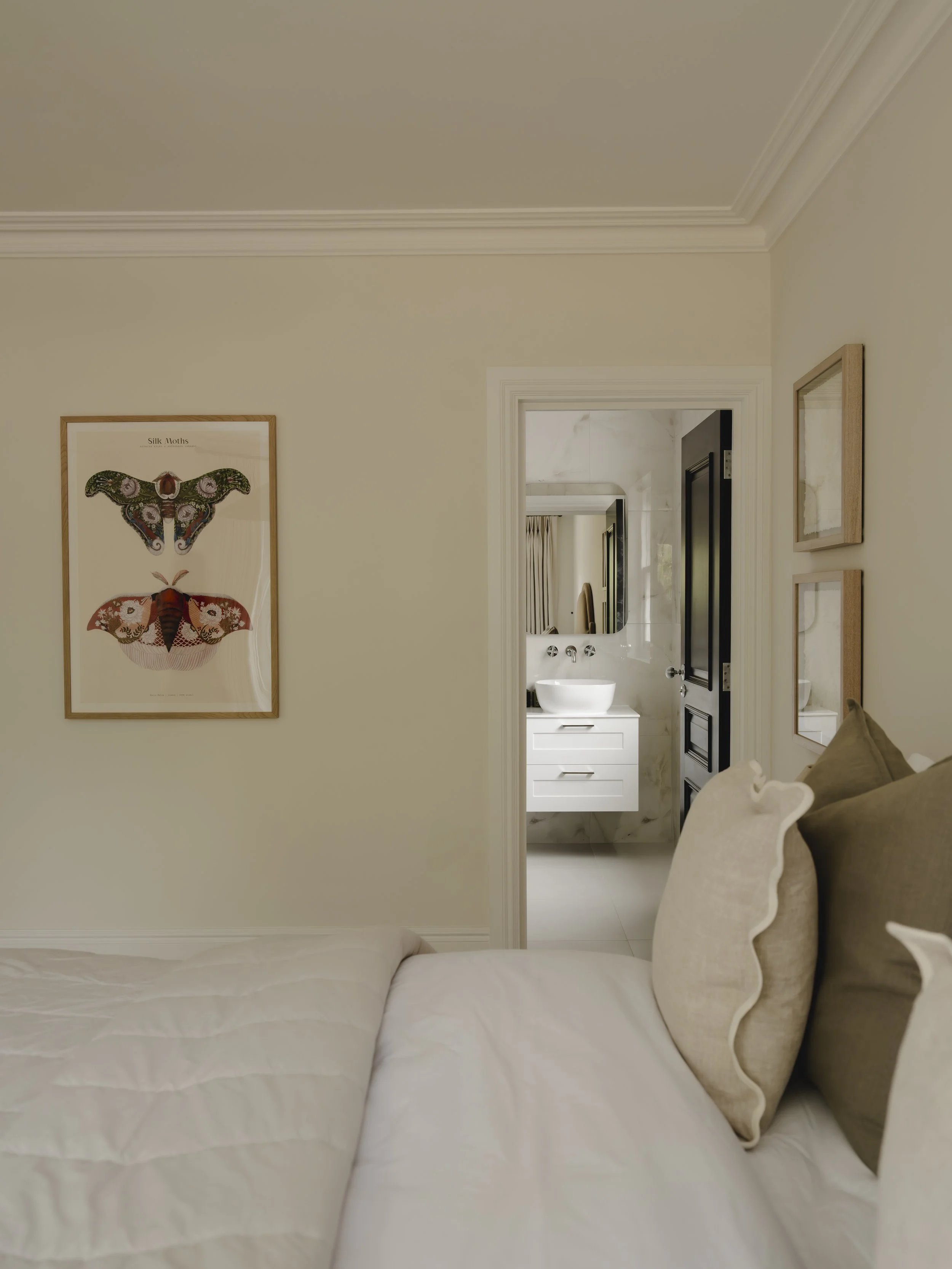 View of a bedroom with a white bed and beige pillows, wall art of two moths, and a doorway leading to a bathroom with white vanity and mirror.
