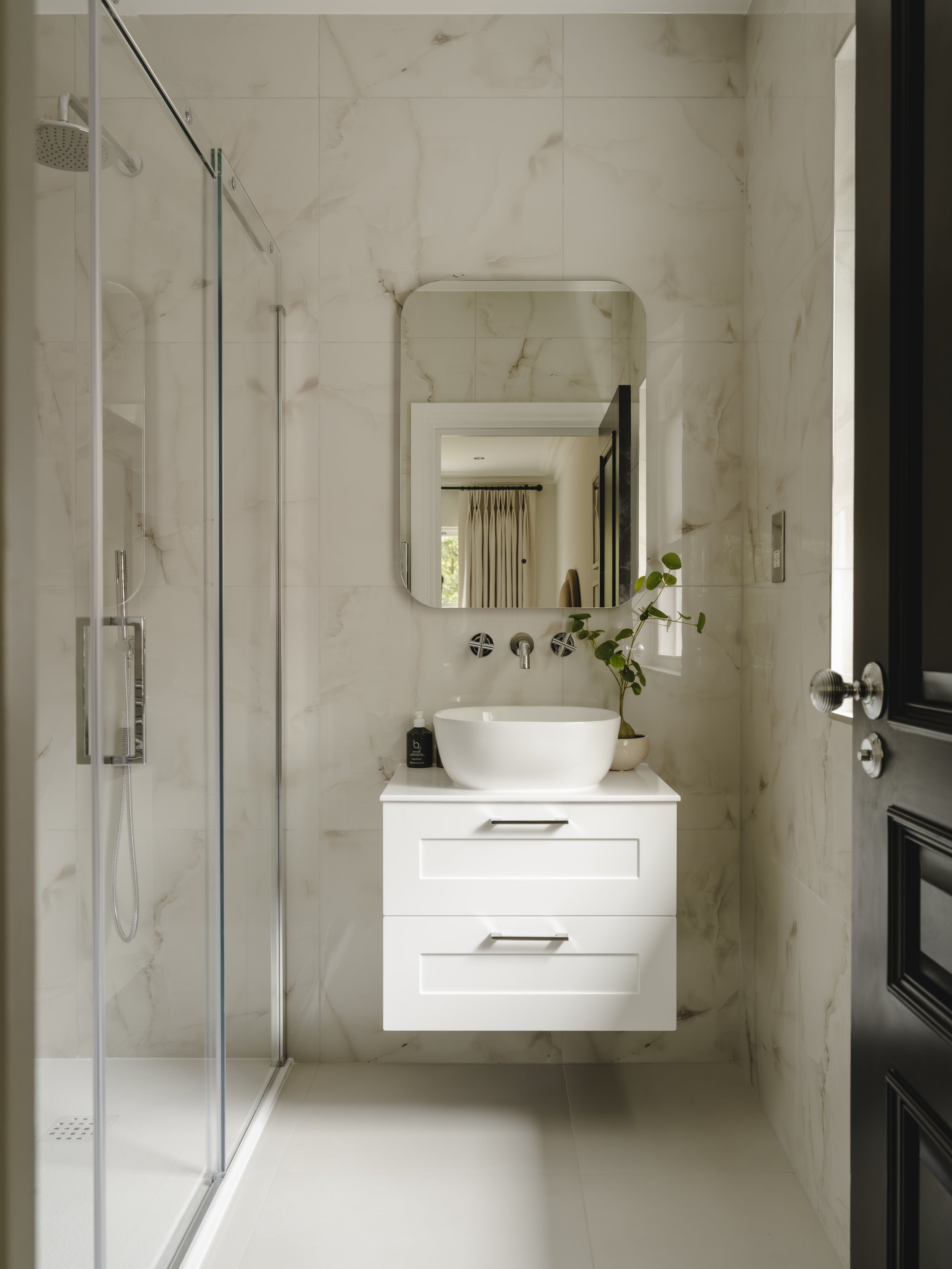 Bathroom with a walk-in shower on the left, a white vanity with a vessel sink, a mirror above, a black soap dispenser, and a potted plant on the right