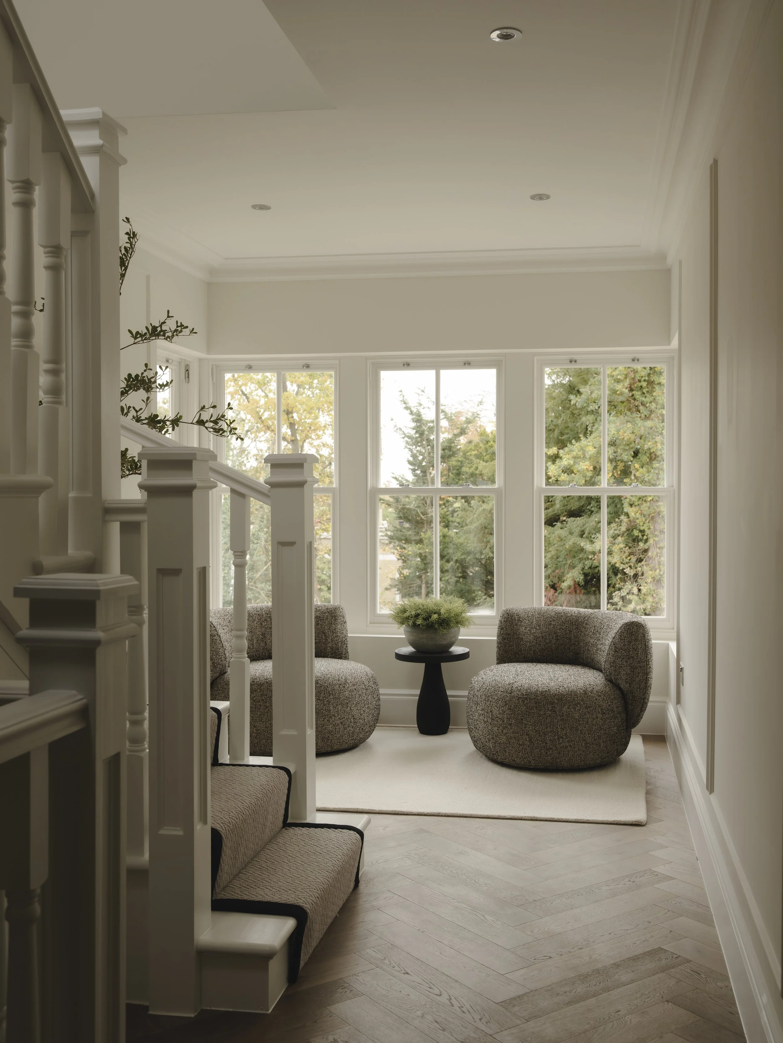 A cozy sitting area with two rounded armchairs, a small black side table with a potted plant, near large windows showing green trees outside, and a stairway on the left.