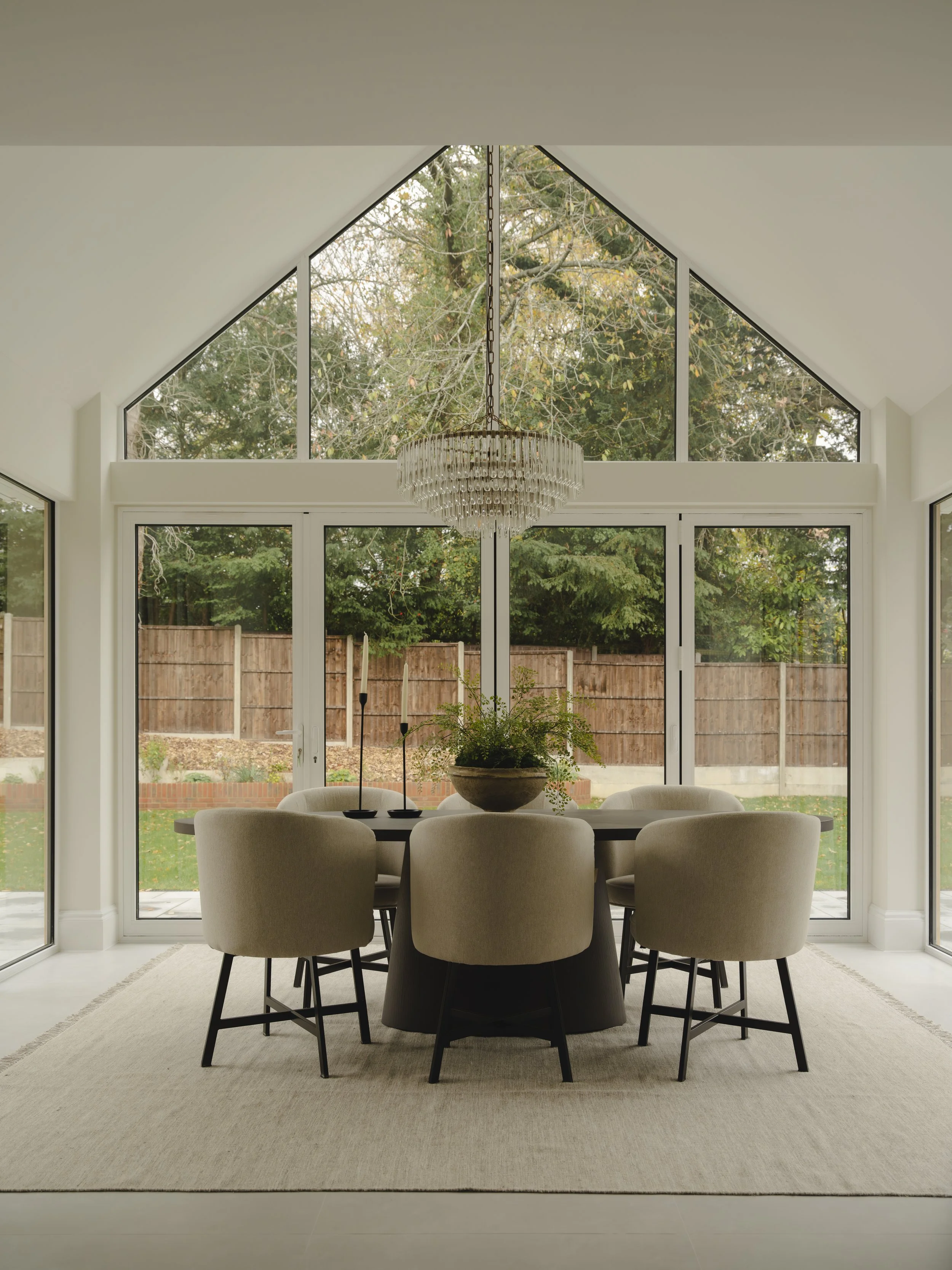 Sunlit dining room with large glass windows and doors, a round table with six beige chairs, a centerpiece plant, and a chandelier hanging from the ceiling.