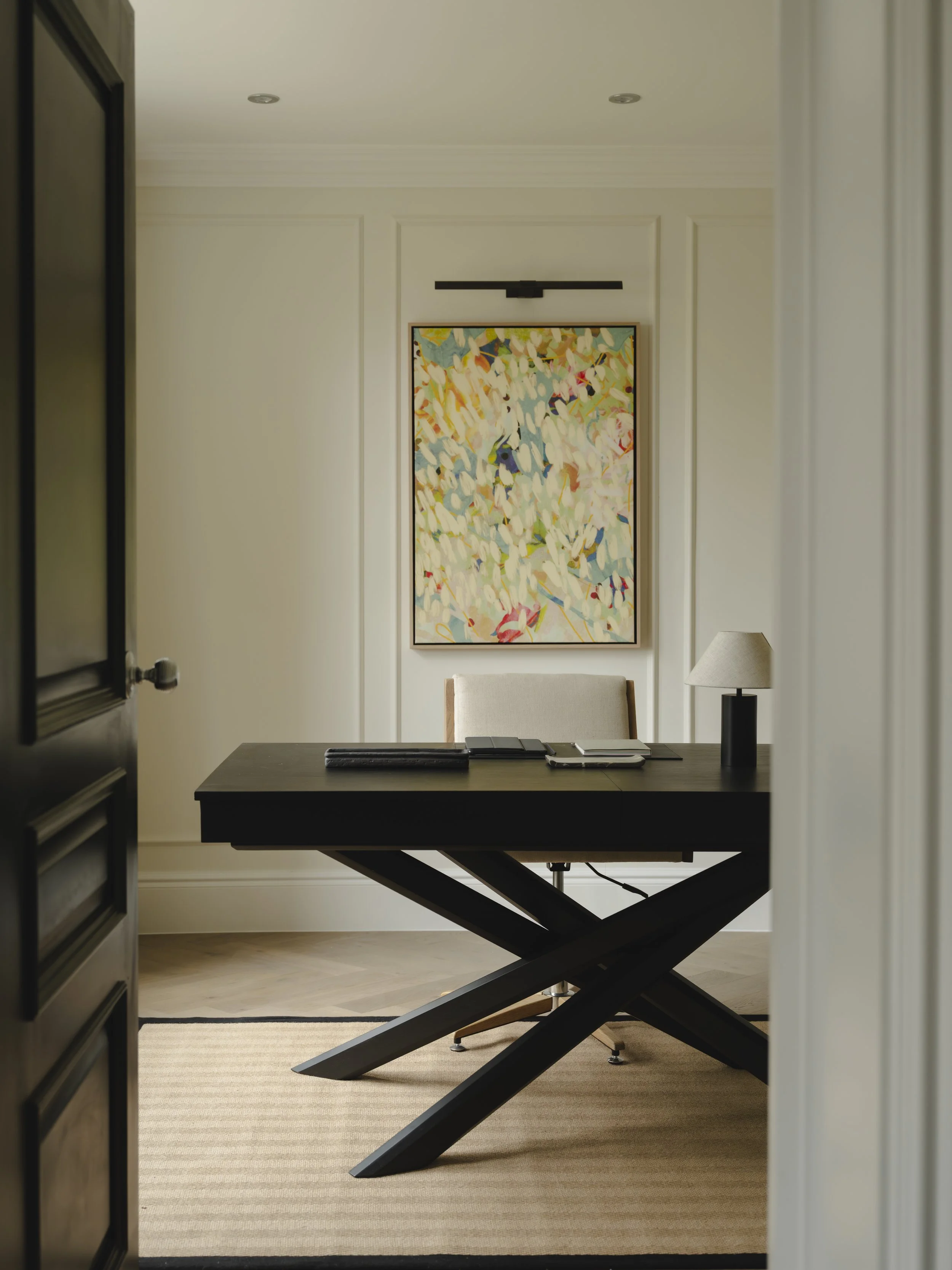 An interior office room with a dark wooden desk, a beige chair, a table lamp, and a colorful abstract painting on the wall.