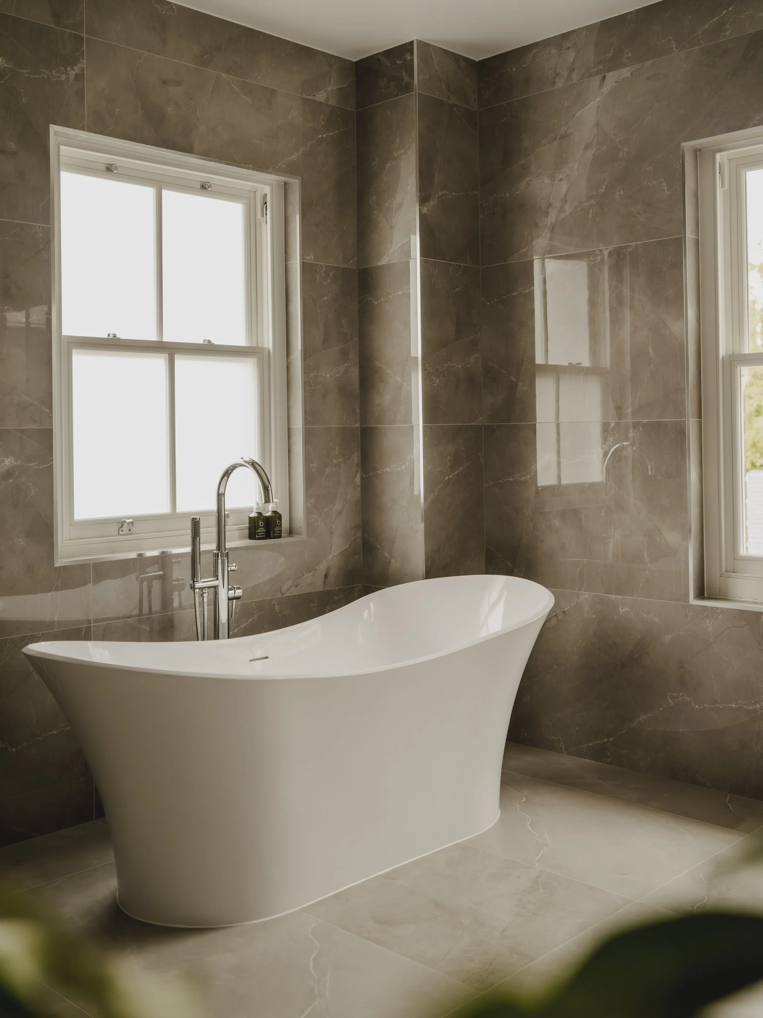 Modern bathroom with a white bathtub, marble walls, and two windows allowing natural light.