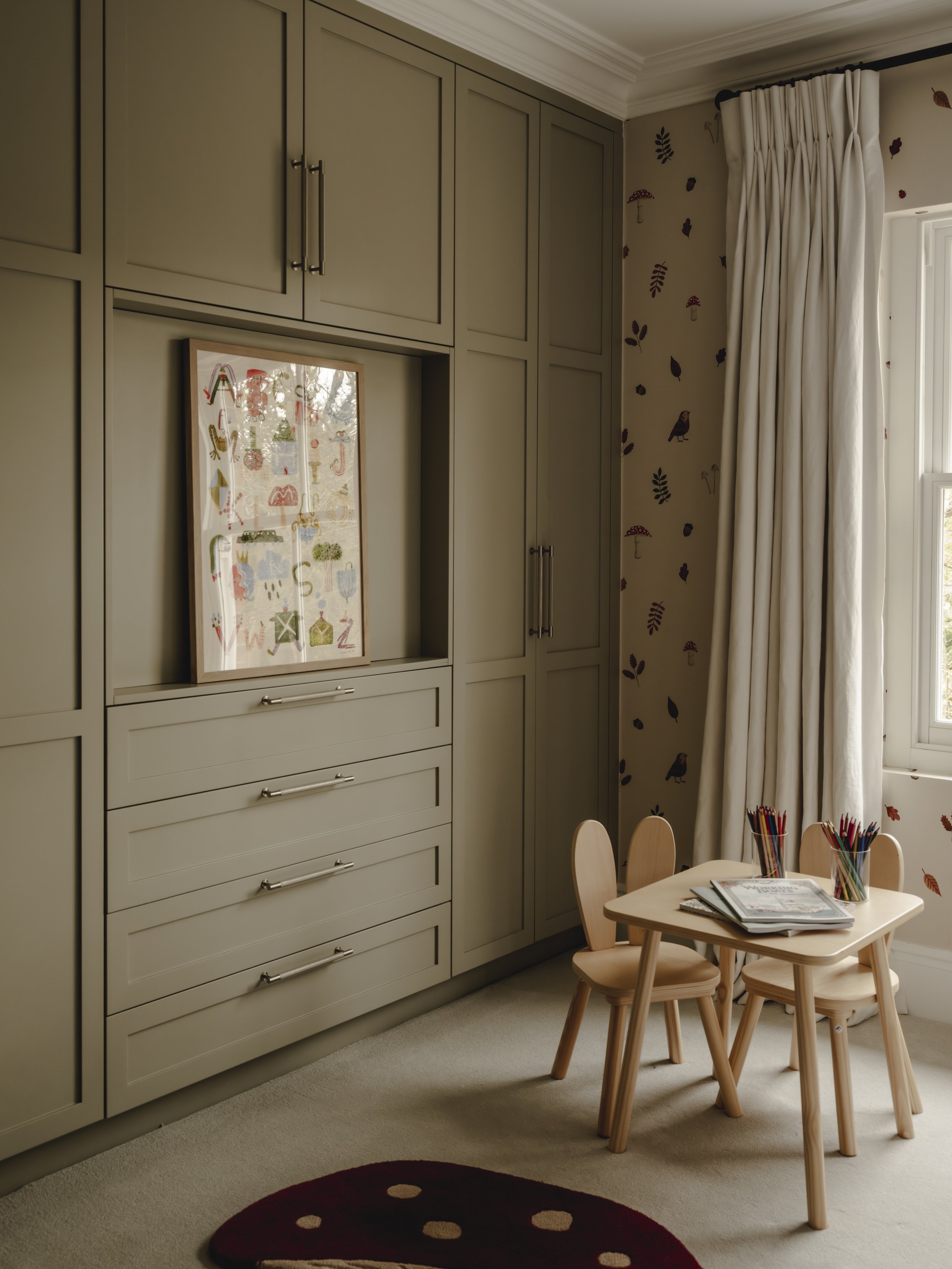 A cozy corner of a child's room with light wooden table and chairs, a large window with white curtains, a wall with patterned wallpaper, and built-in gray cabinetry with a framed colorful artwork.