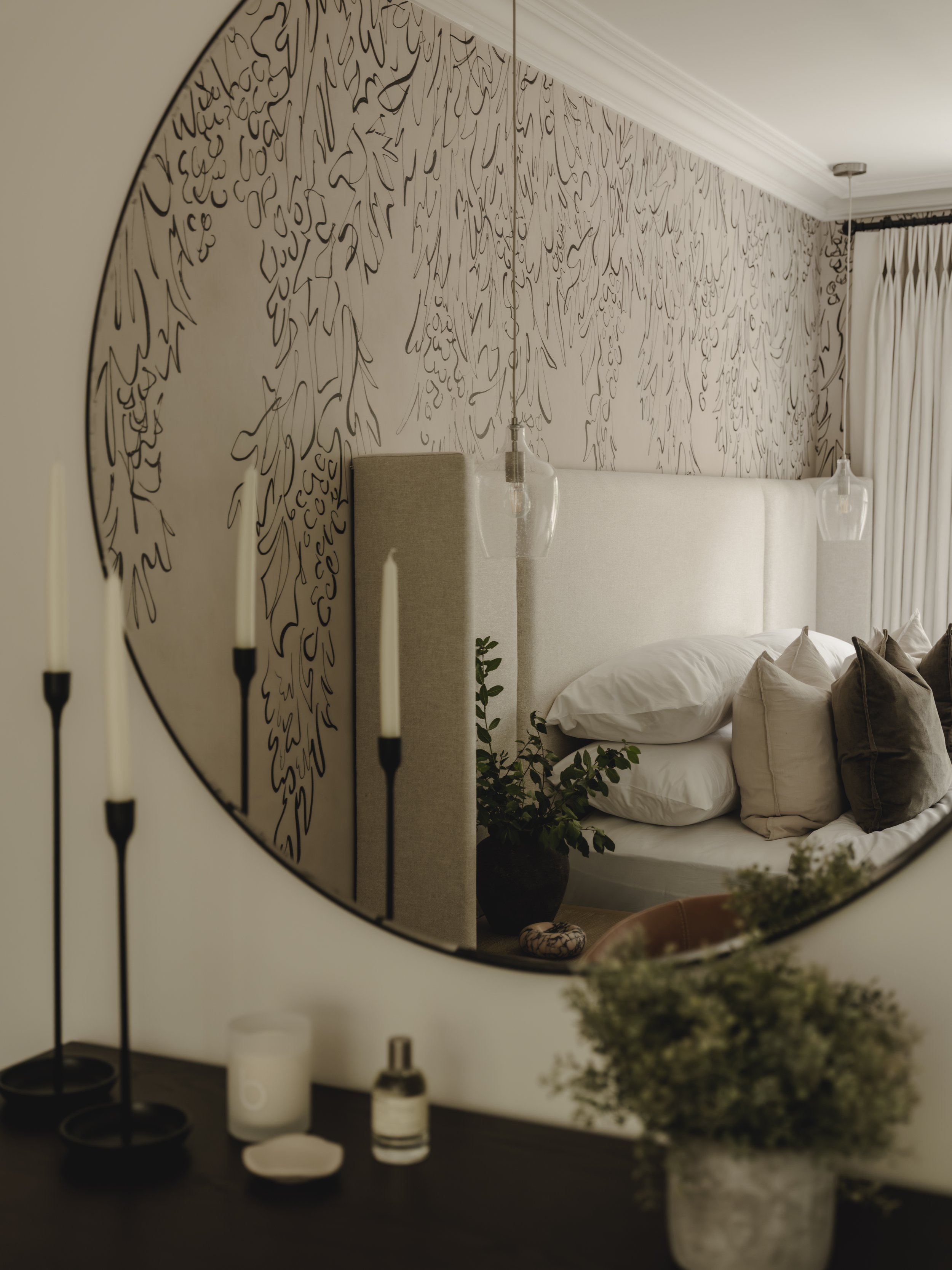 Interior of a bedroom reflected in a round mirror, showing a bed with pillows, a plant, and hanging pendant lights.