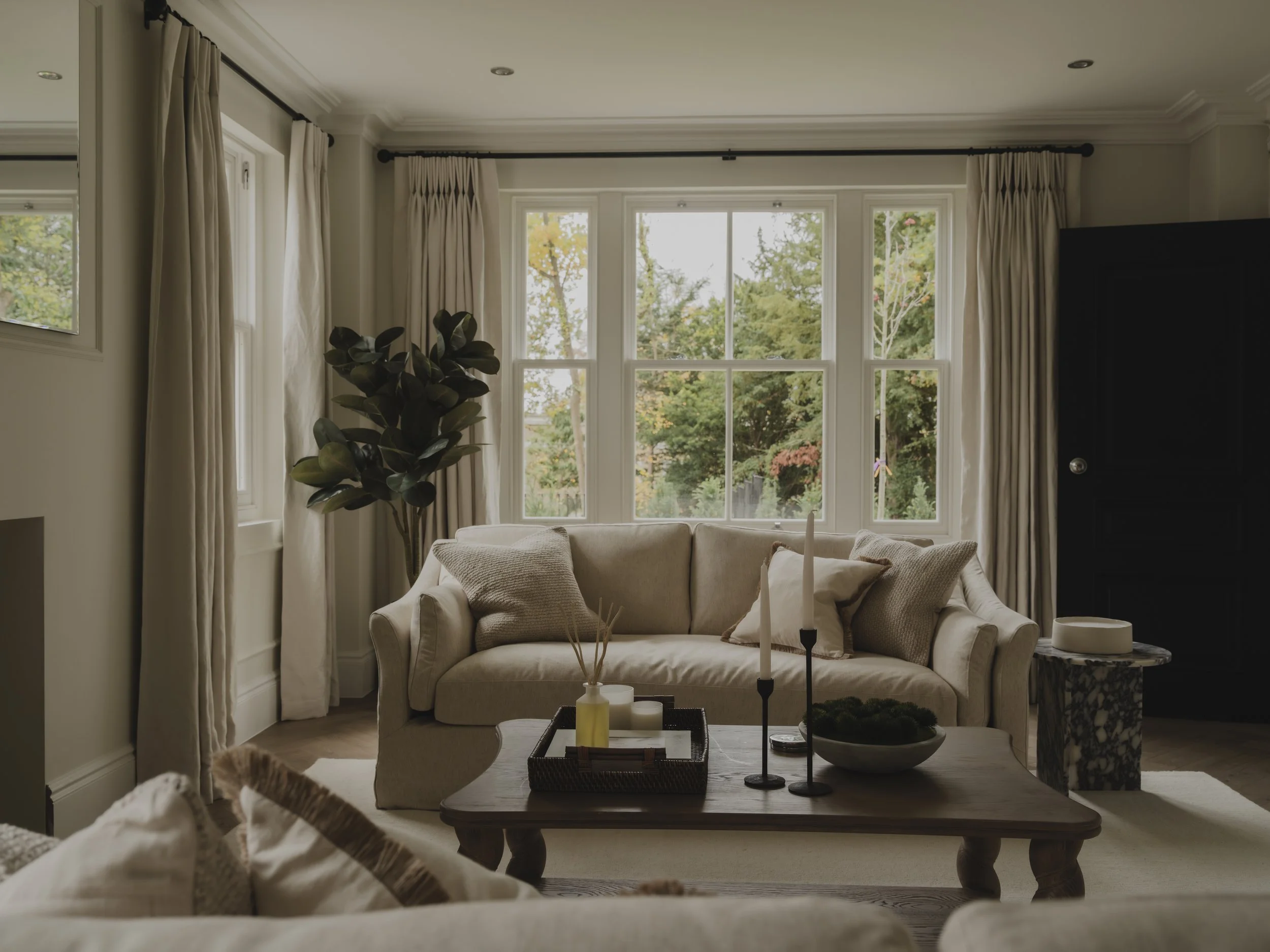 A cozy living room with large windows, a beige sofa with pillows, a wooden coffee table with decorative items, a marble side table, and a tall indoor plant, with a view of trees outside.