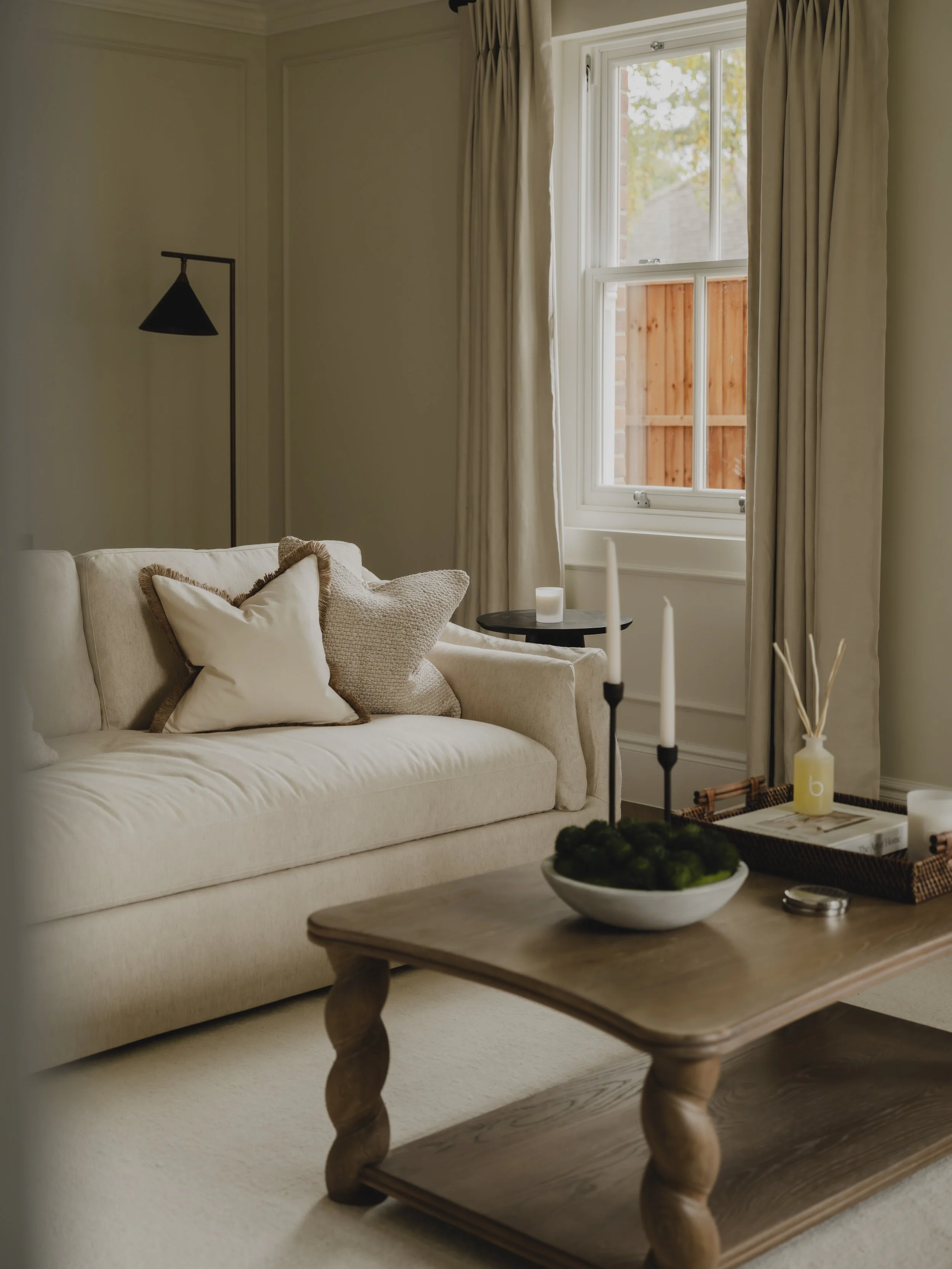 A cozy living room with a cream-colored sofa decorated with beige and white pillows, a wooden coffee table with twisted legs holding a bowl of moss and candles, and a window with cream curtains showing a wooden fence outside.