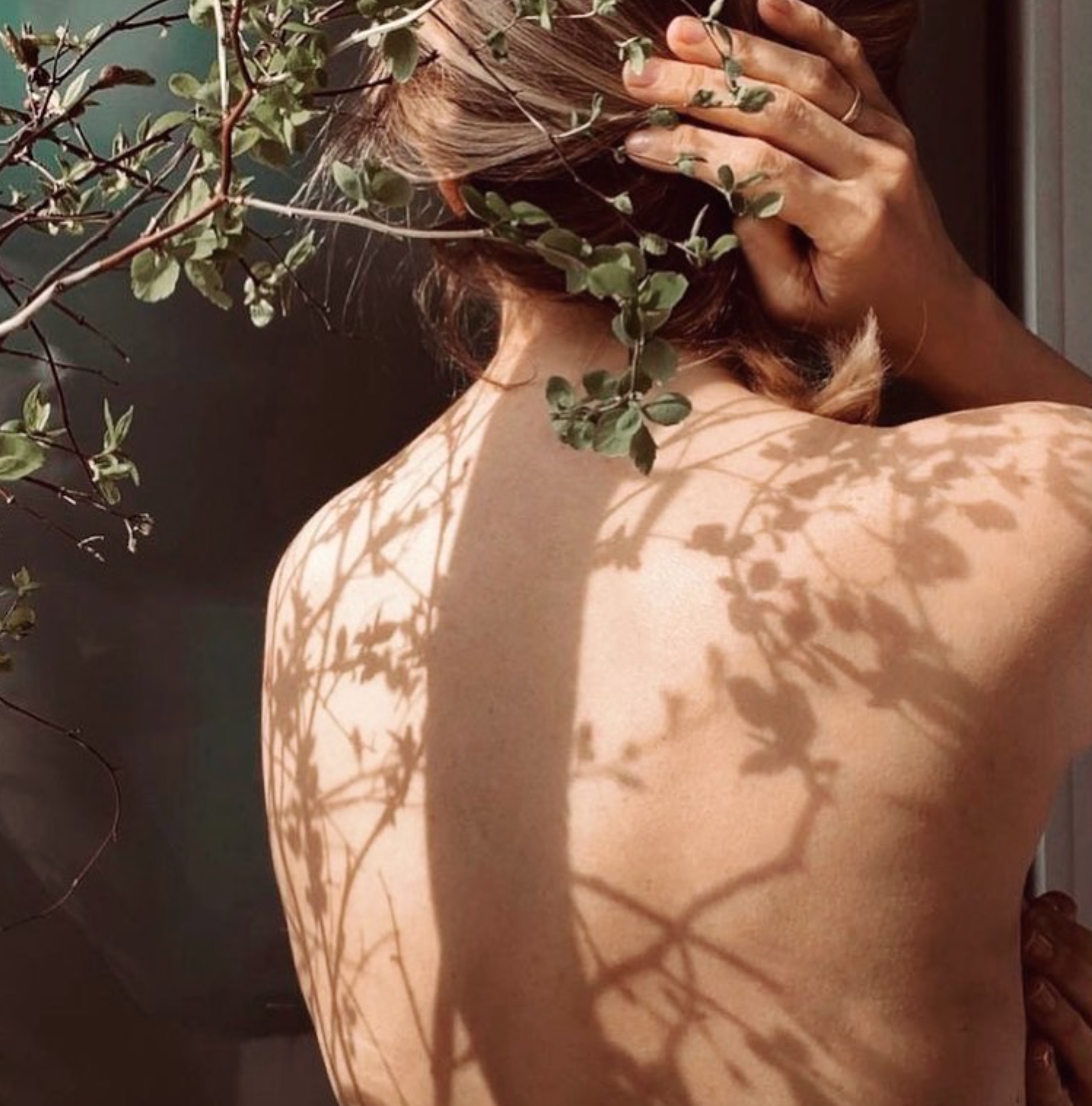 A person with shoulder-length hair covering their face, touching their head with one hand, with shadows of leaves and branches cast on their back and shoulder, and some greenery partially obscures their face and hair.