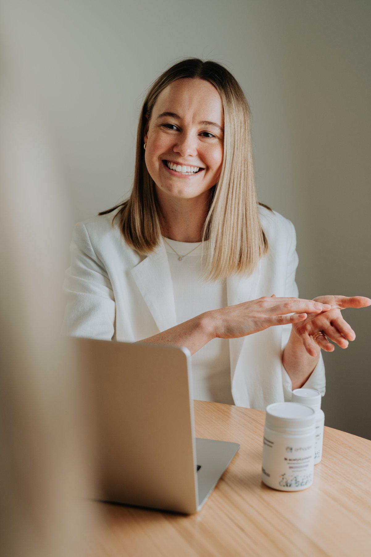 A woman smiling and gesturing with her hands during a discussion, sitting at a desk with a laptop and supplement bottles.