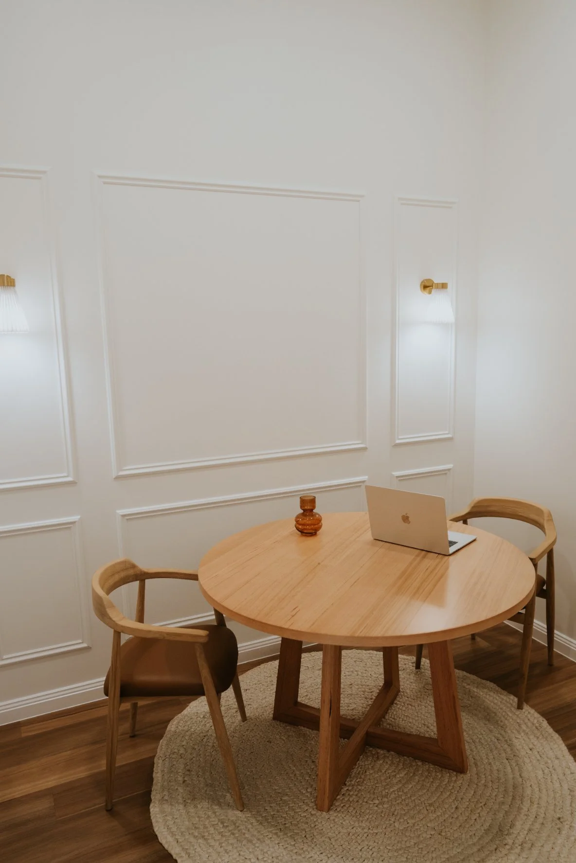 A round wooden table with a laptop and a small decorative item, surrounded by two wooden chairs, in a room with white paneled walls and wall-mounted sconces, on a circular woven rug.