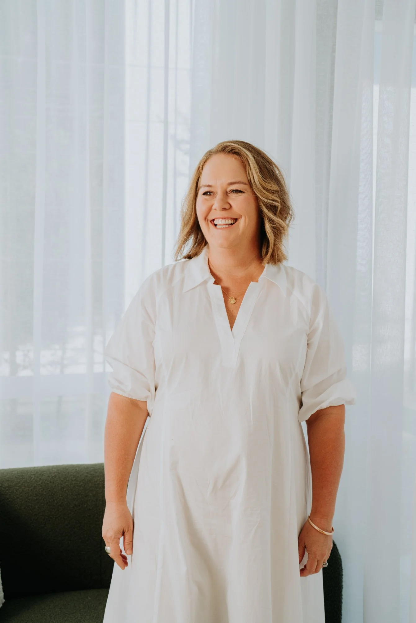 A woman with shoulder-length blonde hair smiling, wearing a white dress, standing in front of a sheer curtain.