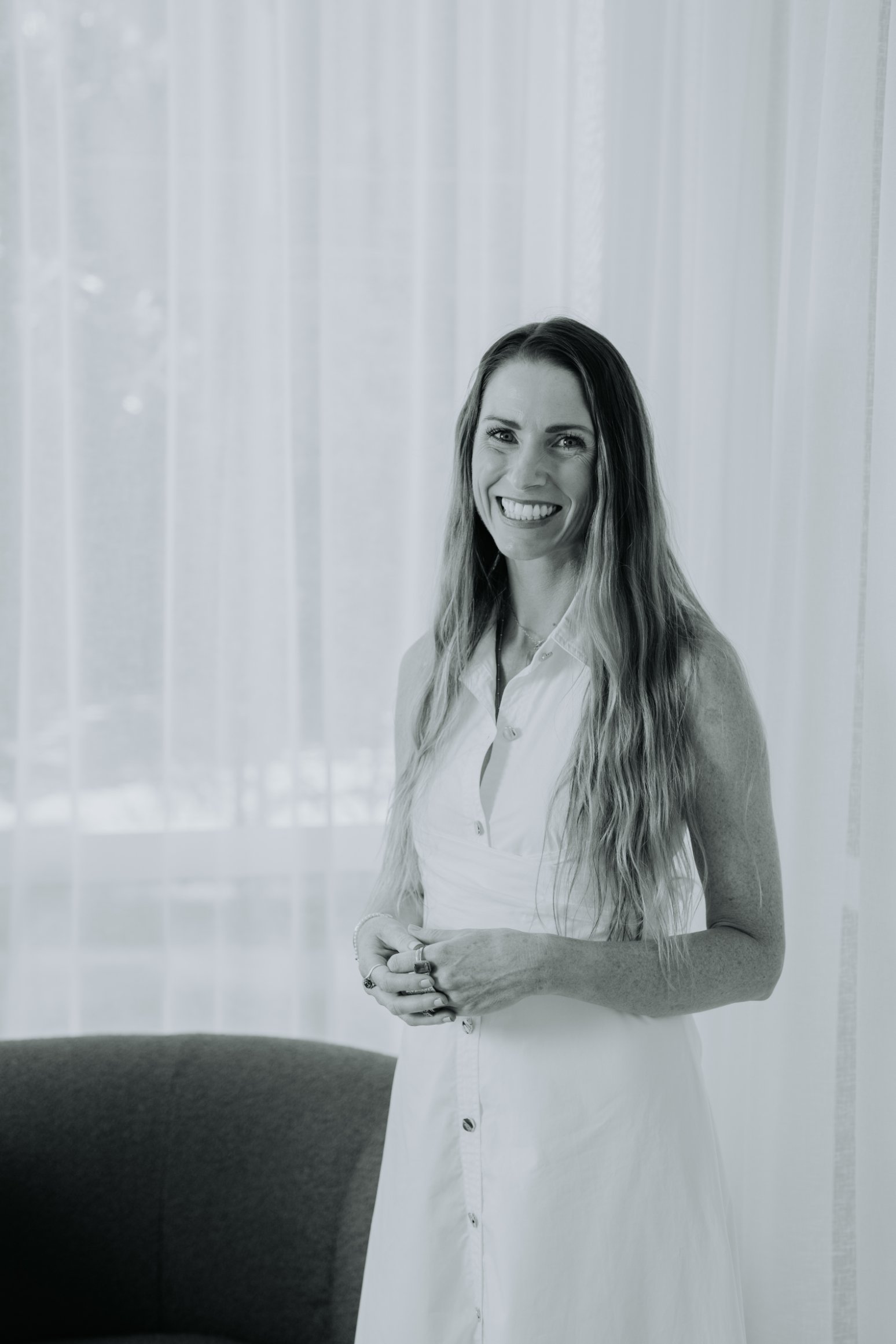 A woman with long wavy hair wearing a sleeveless white button-up dress, smiling and standing in front of sheer curtains with sunlight filtering through.