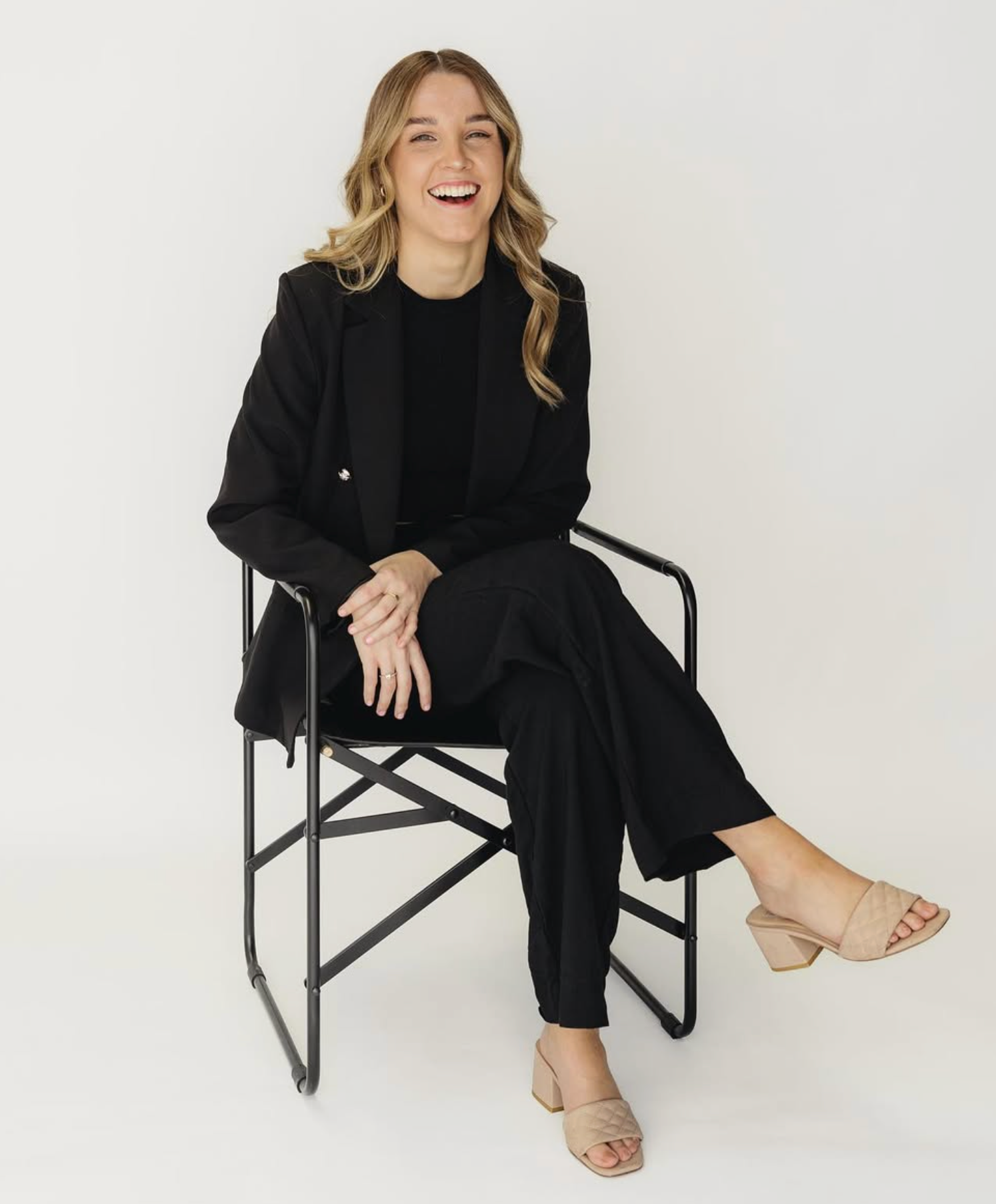 A woman with blonde hair, wearing a black blazer, black pants, beige heeled sandals, is sitting on a black folding chair, smiling, against a plain white background.