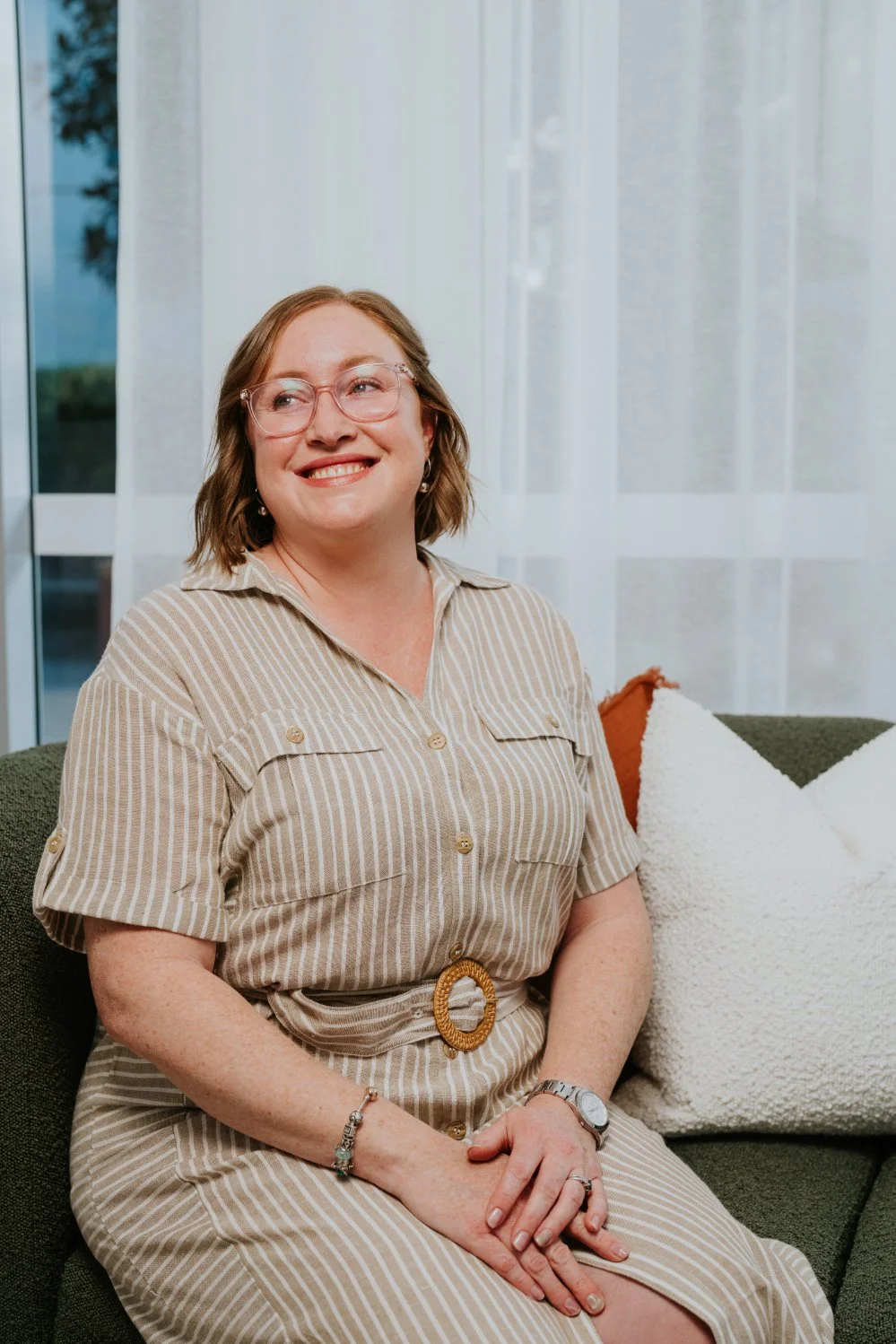 A smiling woman with glasses, wearing a beige striped dress with a belt, sitting on a green couch with white and colored pillows, in front of a window with white curtains.