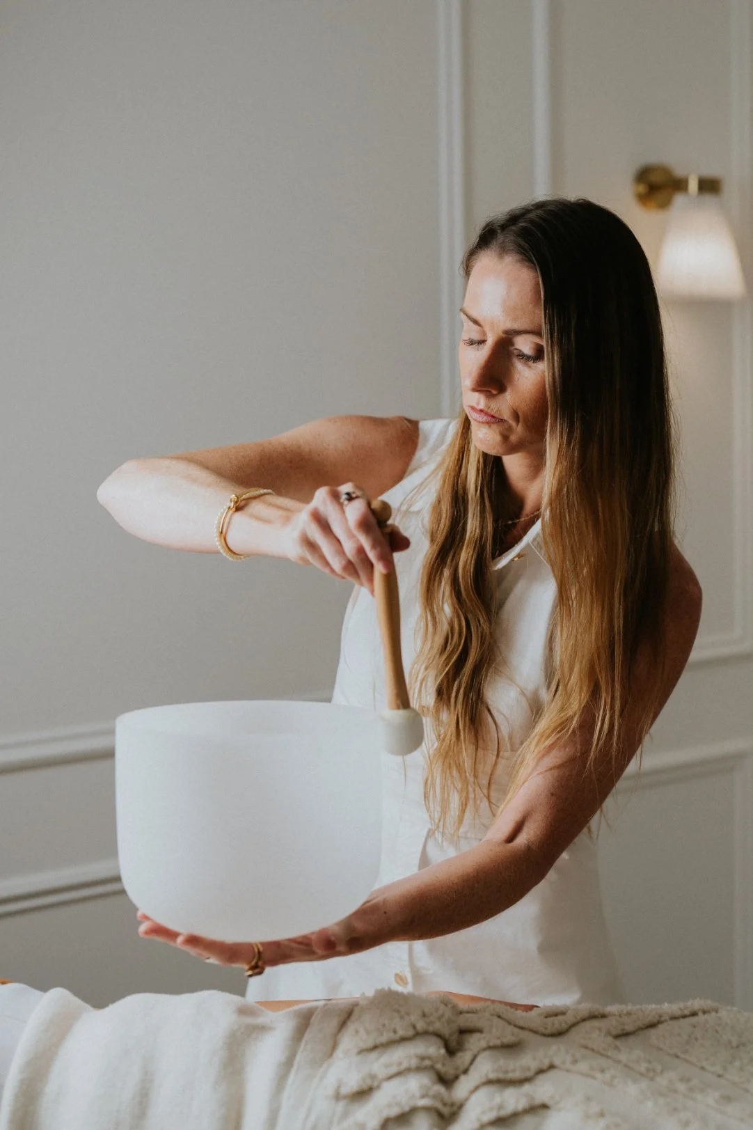 A woman with long brown hair, wearing a white sleeveless top, is holding a singing bowl and a mallet, preparing to strike it. She is standing in a softly lit room with a wall-mounted lamp in the background.
