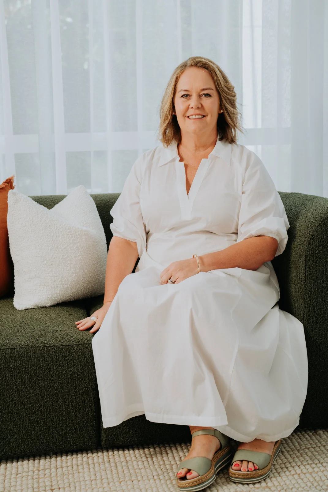 A woman with shoulder-length blonde hair, wearing a white dress, sitting on a dark green sofa with cream pillows, in front of a sheer white curtain with a window behind.