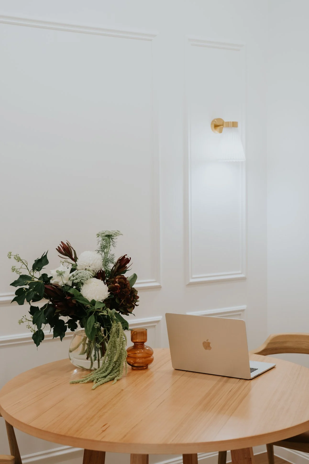 A wooden round table with a bouquet of flowers in a glass vase, a small amber-colored vase, and a closed silver MacBook laptop in a room with white paneled walls and a wall-mounted light fixture.