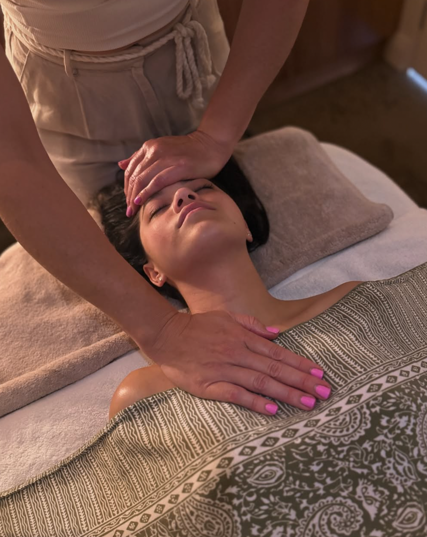 A woman receiving a facial massage from a massage therapist, lying on a massage table with her eyes closed.