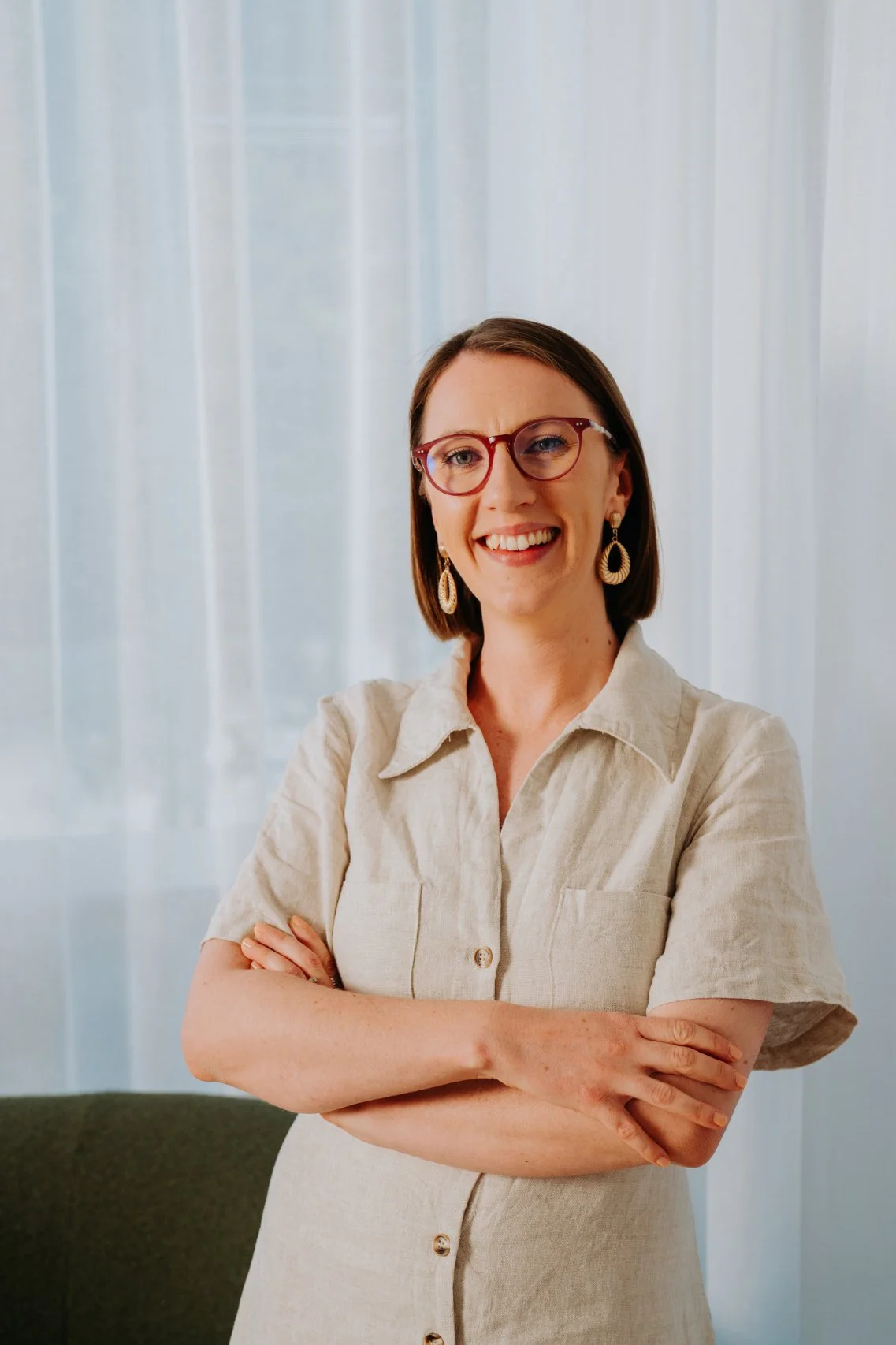 A woman with short brown hair, wearing red glasses, gold earrings, and a beige button-up shirt, smiling with her arms crossed in front of a light-colored, semi-transparent curtain.