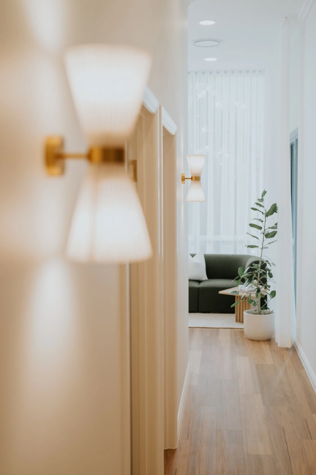 Hallway with wall-mounted sconces leading to a waiting area with a green sofa, a round wooden side table, a potted plant, sheer curtains, and natural light.
