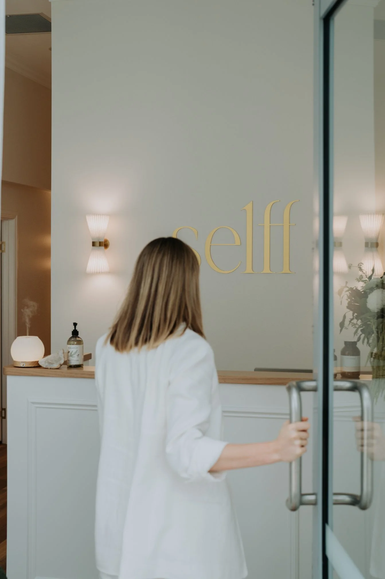 A woman in a white blazer opening a glass door into a wellness or spa center with the word 'self' on the wall.
