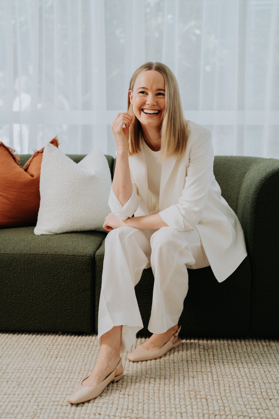 A woman with blonde hair wearing a white suit and beige heels, sitting on a green sofa with decorative pillows, smiling and laughing in a well-lit room with sheer curtains in the background.