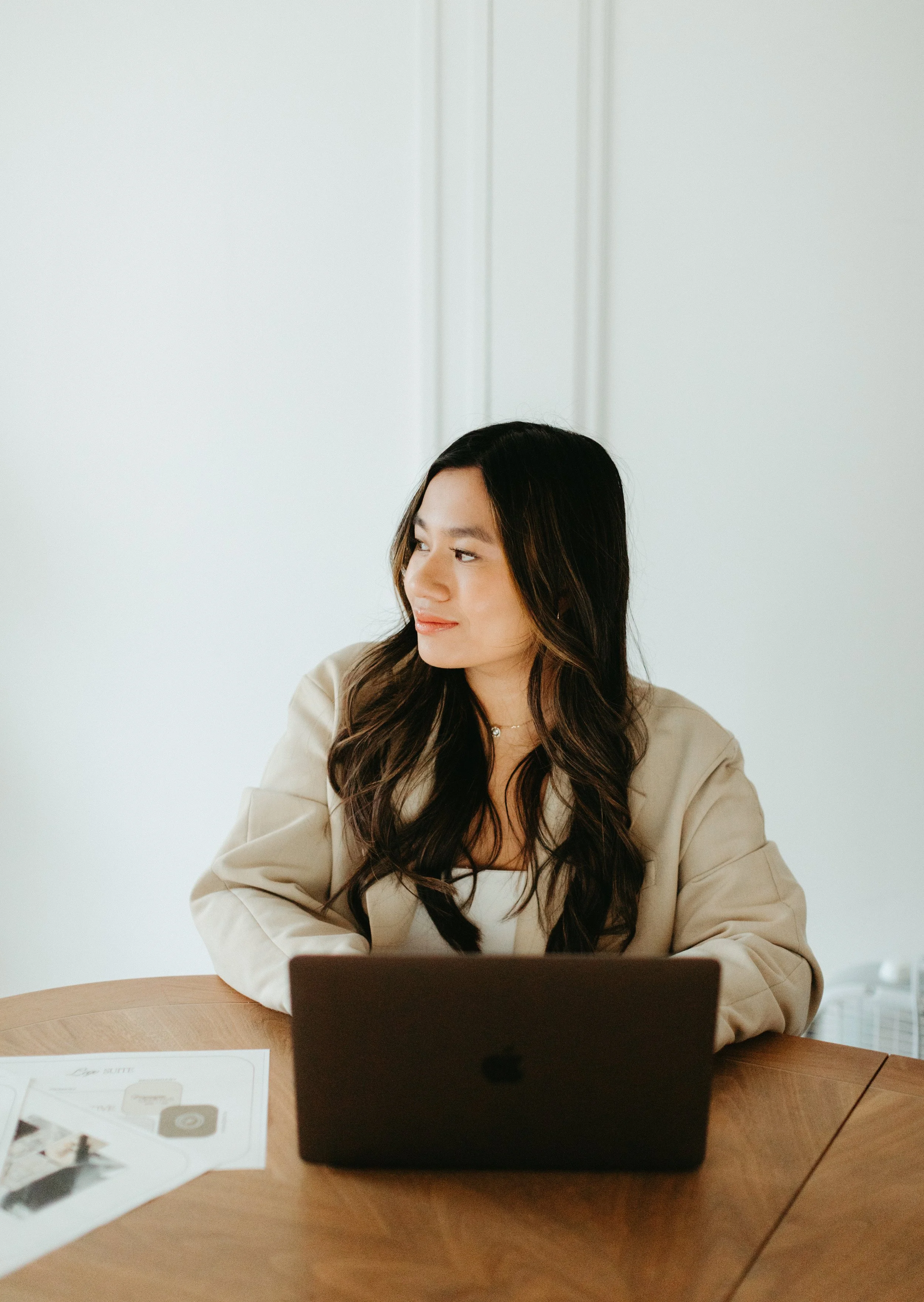 A woman with long dark hair sitting at a wooden table using a laptop, wearing a beige blazer and white top, with a white wall in the background.