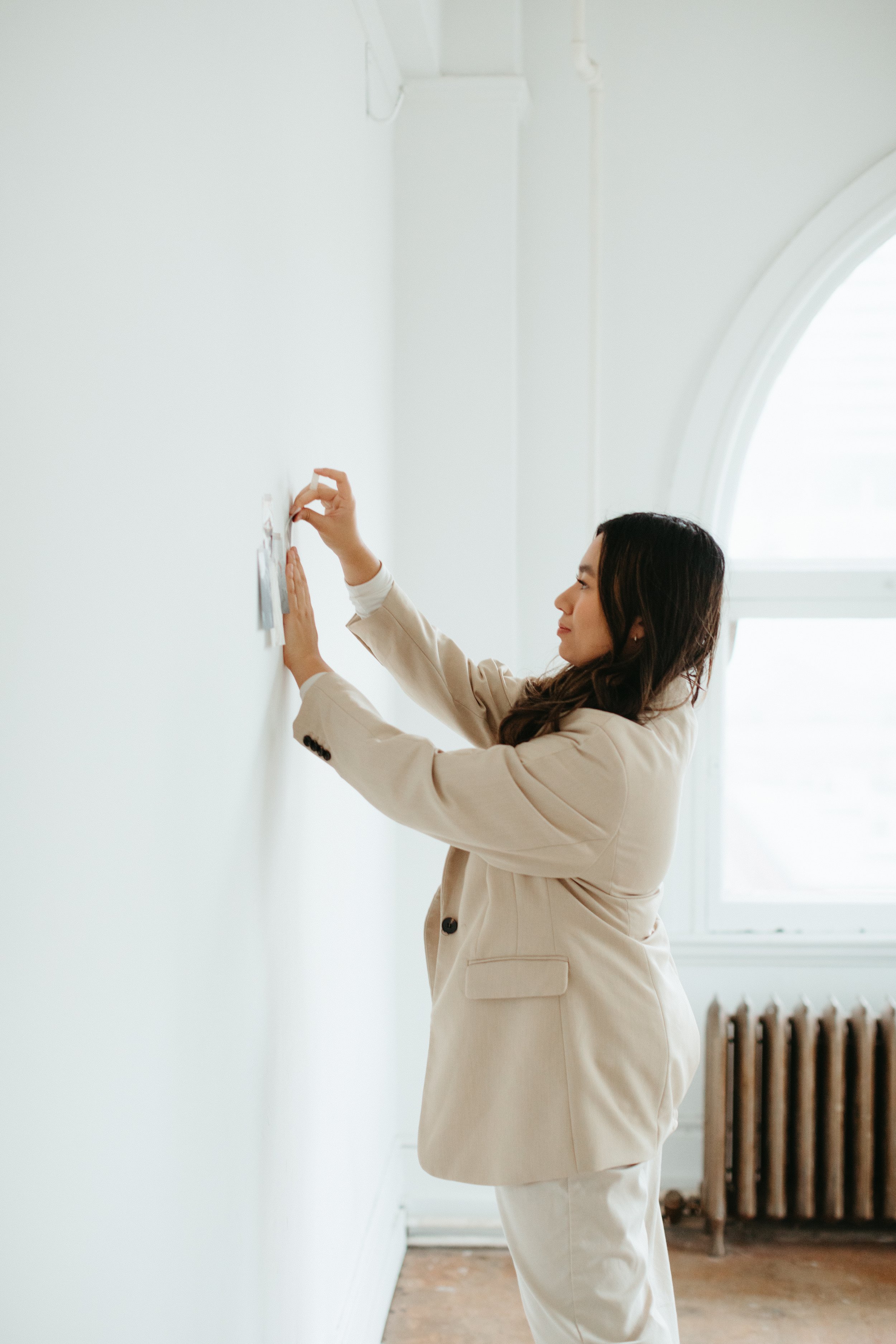 Woman putting up photos or notes on a white wall in a bright room with large window.