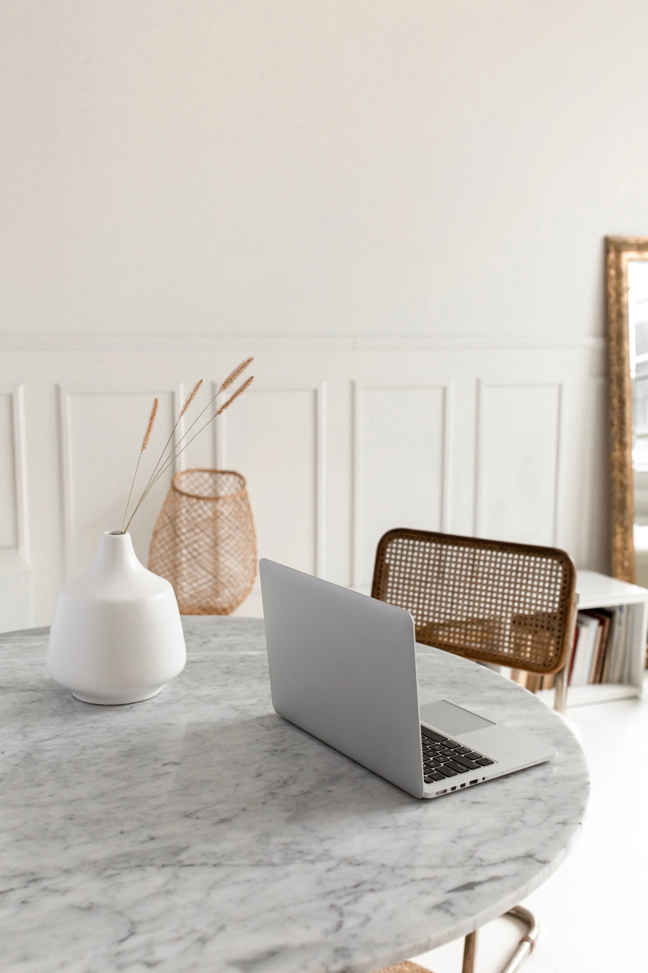 Minimalist workspace with open laptop, white vase with dried grass, wicker basket, and wooden chair in a bright room.