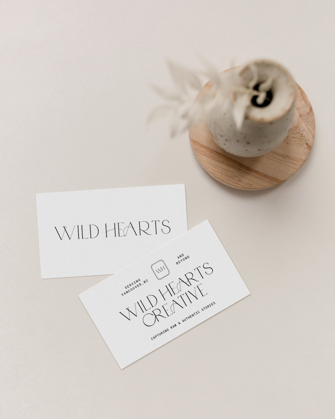 Business cards for Wild Hearts Creative with the tagline "Capturing Raw & Authentic Stories", placed on a white surface next to a small round wooden stand holding a decorative plant in a white vase.