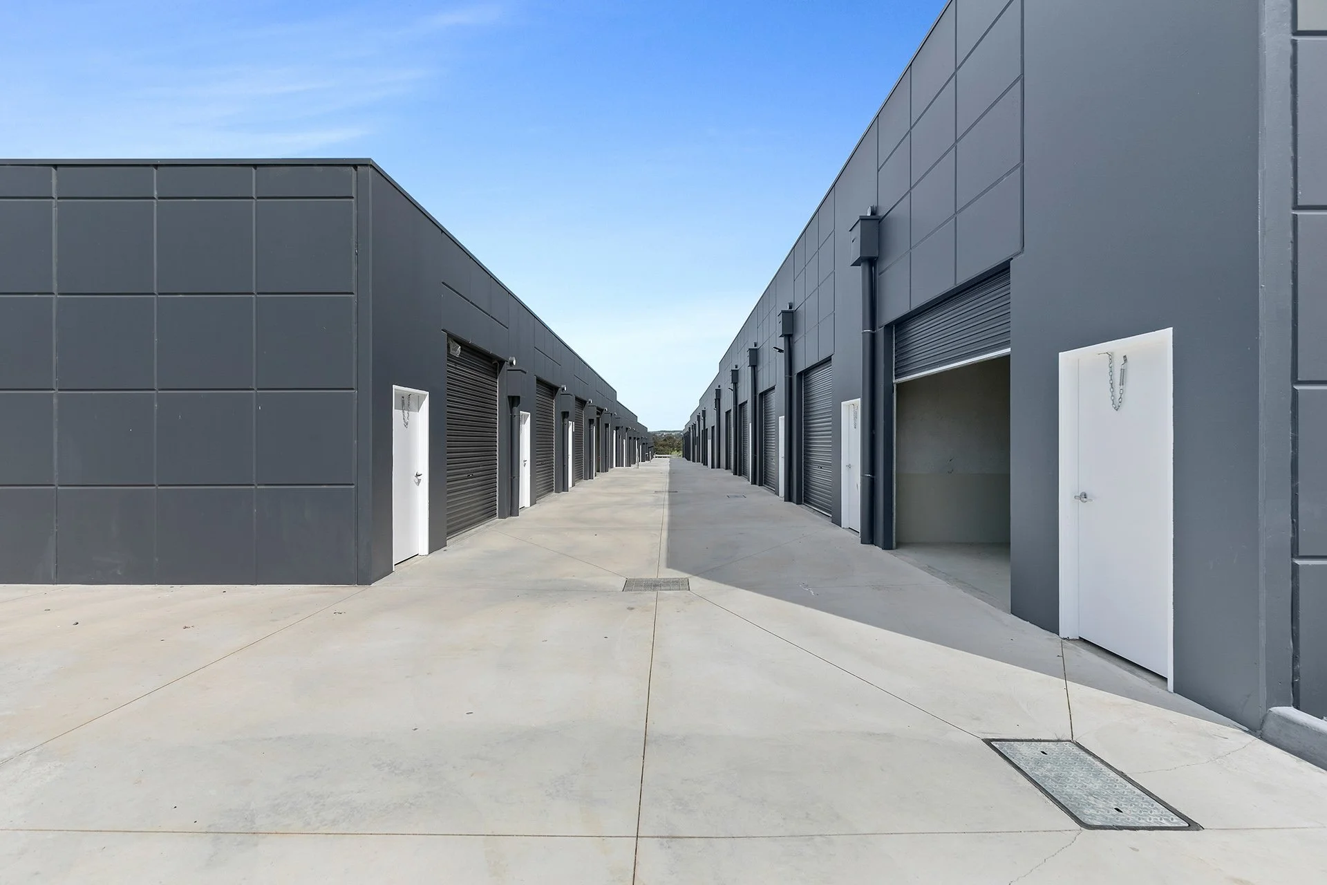 Two parallel rows of gray storage units or garages with closed and open roll-up doors, on a concrete surface under a blue sky.