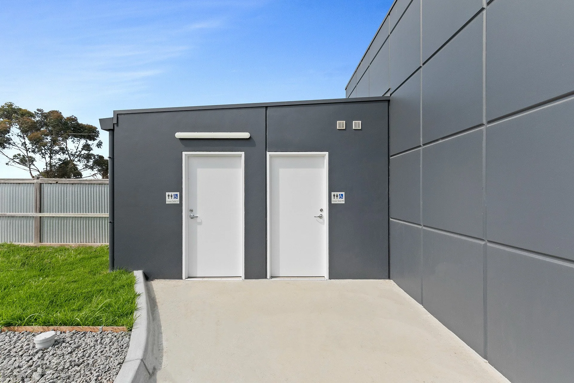 Exterior of a gray building with two white doors, one marked as handicap accessible, on a concrete sidewalk with a small green lawn and a gravel area, clear blue sky in the background.