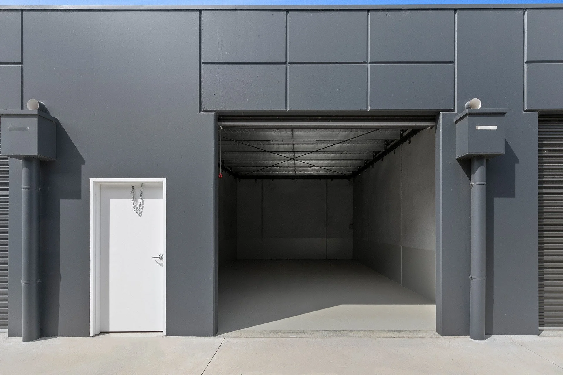 Gray industrial building with a large open garage door and a small white door with a chain, set on a concrete surface.